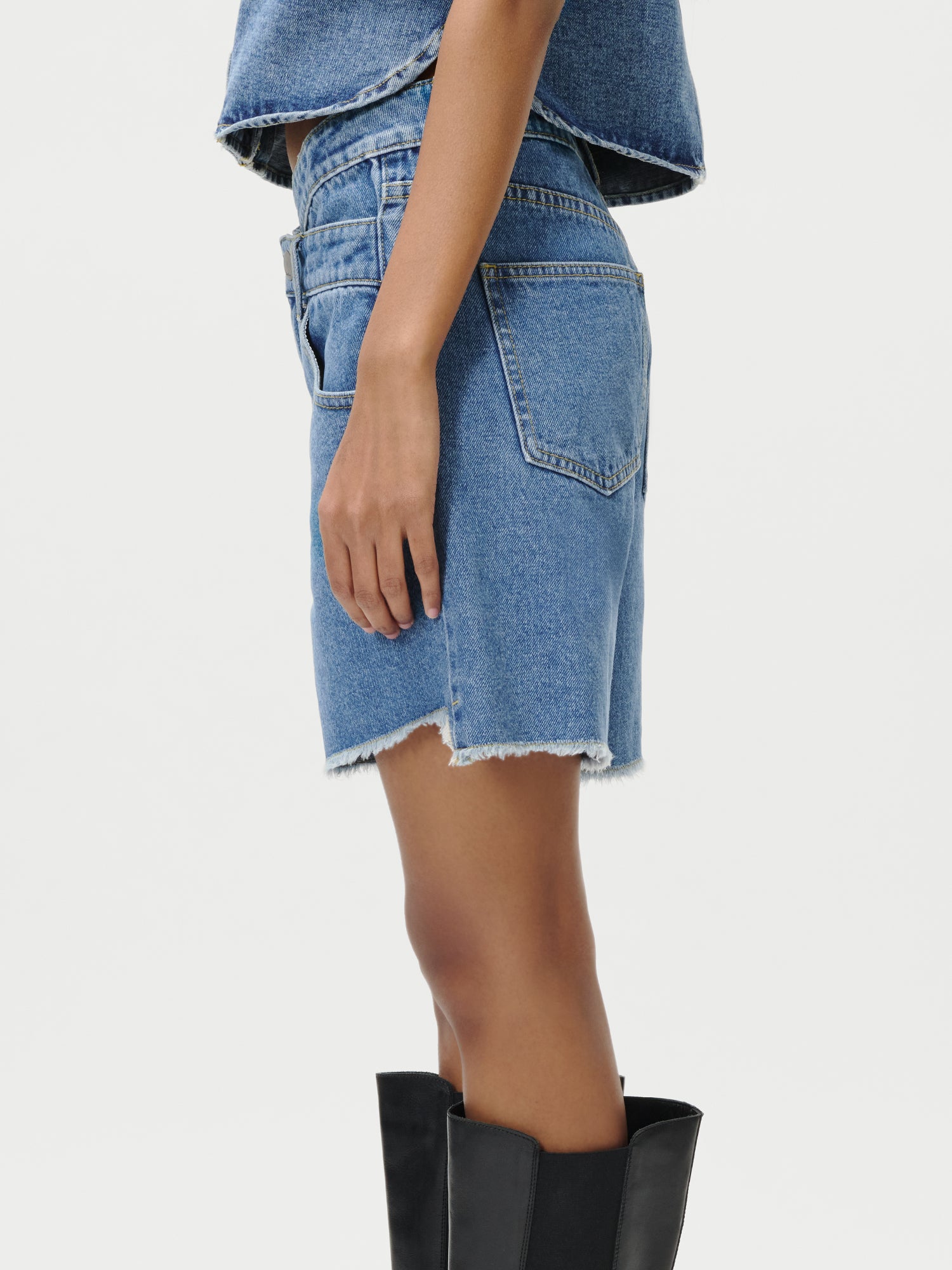 Eine Person im Profil trägt Shirin Shorts von LeGer by Lena Gercke aus blauem Denim mit rohem Saum, ein passendes Denim-Top und schwarze kniehohe Stiefel vor einem schlichten weißen Hintergrund. Größe 36.