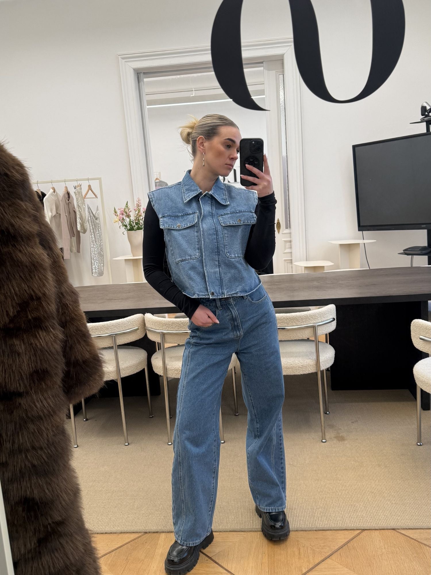 Eine Frau in einem stilvollen Zimmer macht ein Selfie im Spiegel. Sie trägt eine Shirin Pants von LeGer by Lena Gercke in Größe 36, eine Jeansweste und ein schwarzes langärmeliges Oberteil; ihr Haar ist zu einem Dutt gebunden. Stühle, Tisch, Fernseher und hängende Kleidung erscheinen hinter ihr.