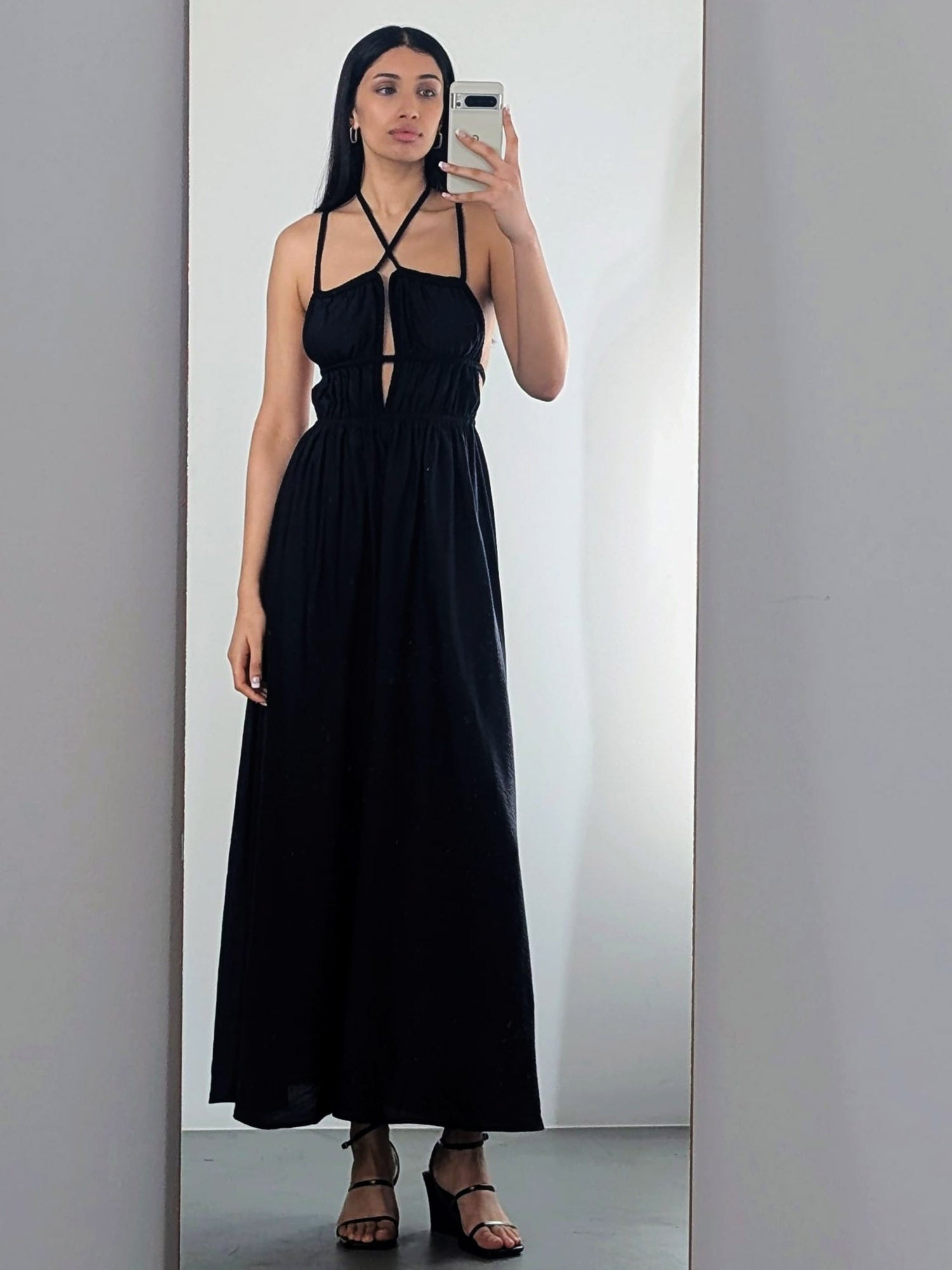 Eine Frau macht ein Spiegel-Selfie in einem LeGer by Lena Gercke Hanke Dress - einem langen schwarzen Neckholder-Kleid mit ausgeschnittenen Details - und schwarzen hochhackigen Sandalen. Der Hintergrund ist minimalistisch und in hellen Farben gehalten.
