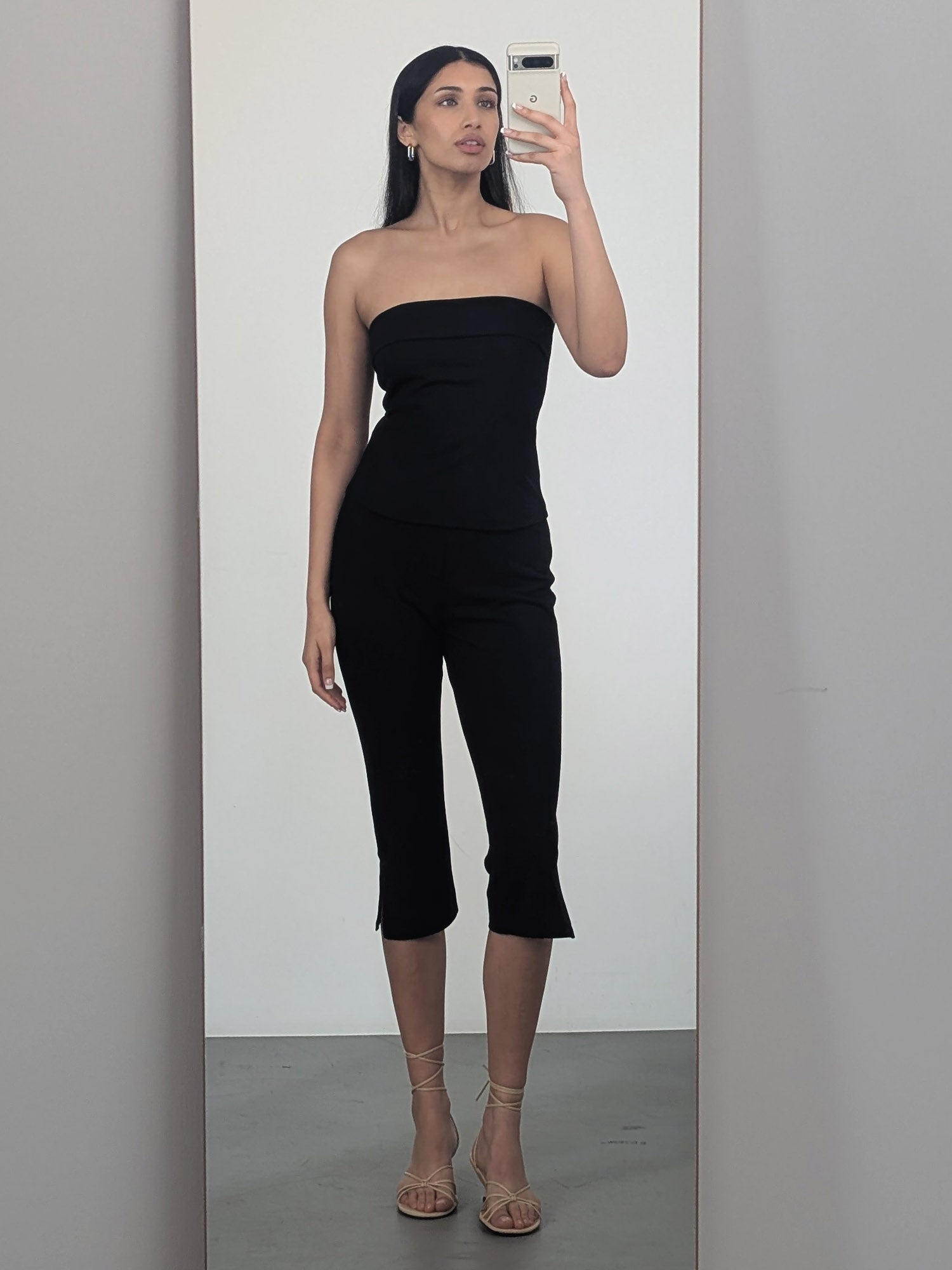 Eine Frau macht ein Spiegel-Selfie in einem trägerlosen schwarzen Top, beigen Riemchensandalen und der Dunja-Hose von LeGer by Lena Gercke und kreiert so einen stylischen Damen-Hosen-Look vor einem minimalistischen Indoor-Hintergrund.