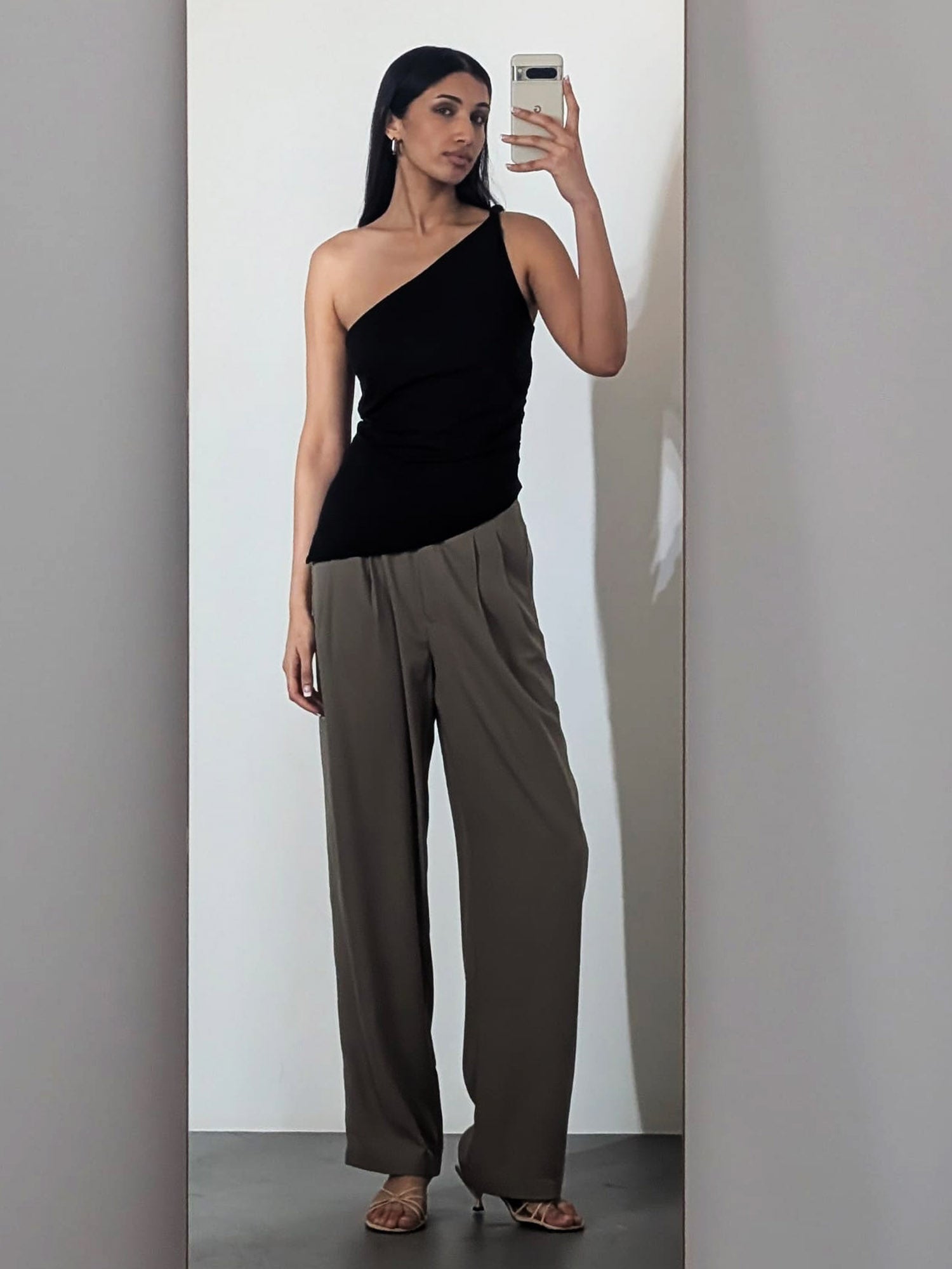 Eine Frau macht ein Selfie mit dem LeGer by Lena Gercke Darleen Top - ein schwarzes One-Shoulder-Top - in Kombination mit einer weiten, hoch taillierten Hose in Taupe und offenen Absätzen vor einem schlichten Hintergrund.