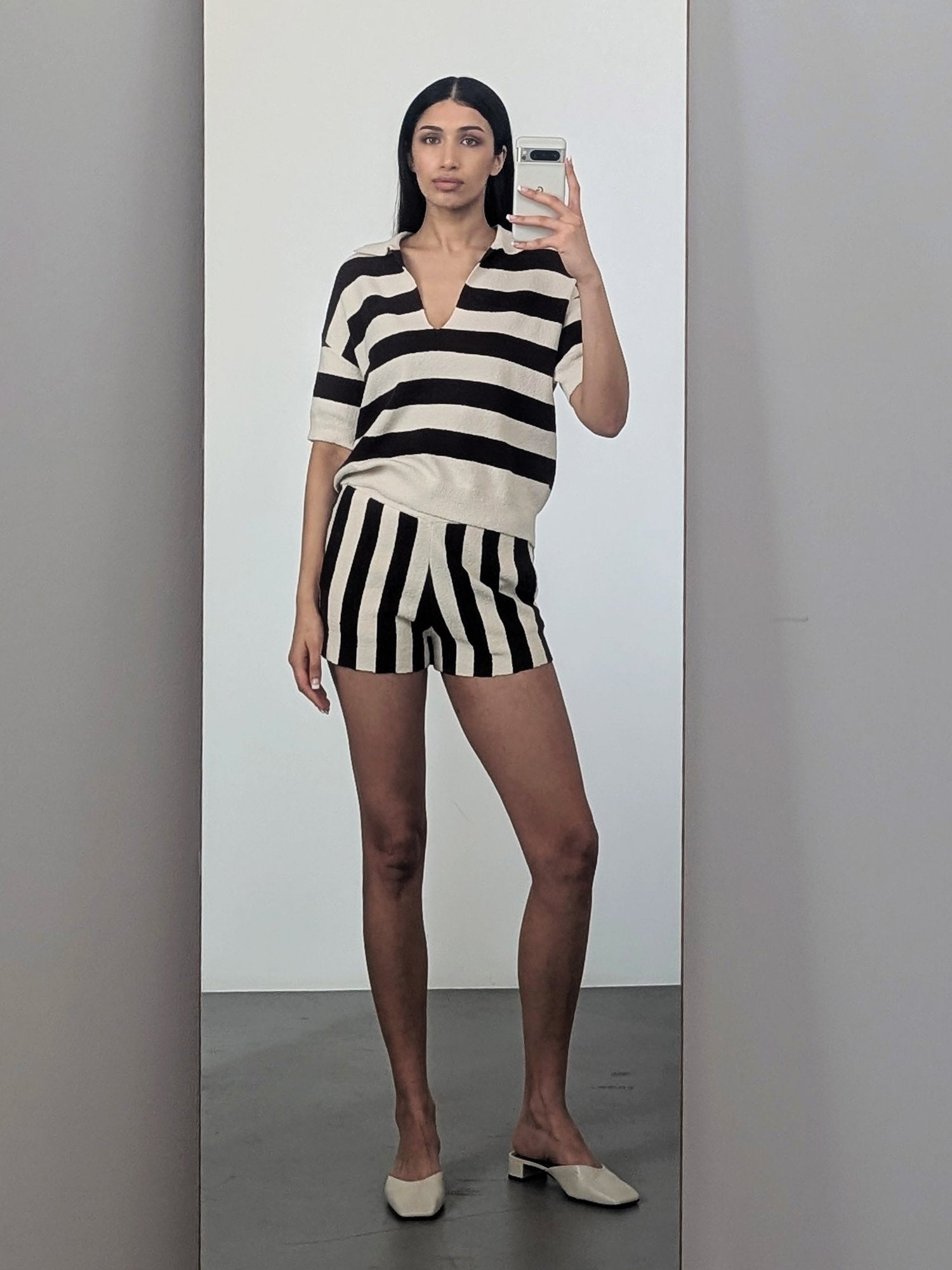 Eine Frau macht ein lässiges Selfie im Spiegel. Sie trägt das schwarz-weiß gestreifte Lisey T-Shirt von LeGer by Lena Gercke mit passenden Shorts, drinnen auf einem grauen Boden mit neutralen Wänden und cremefarbenen Slippern.