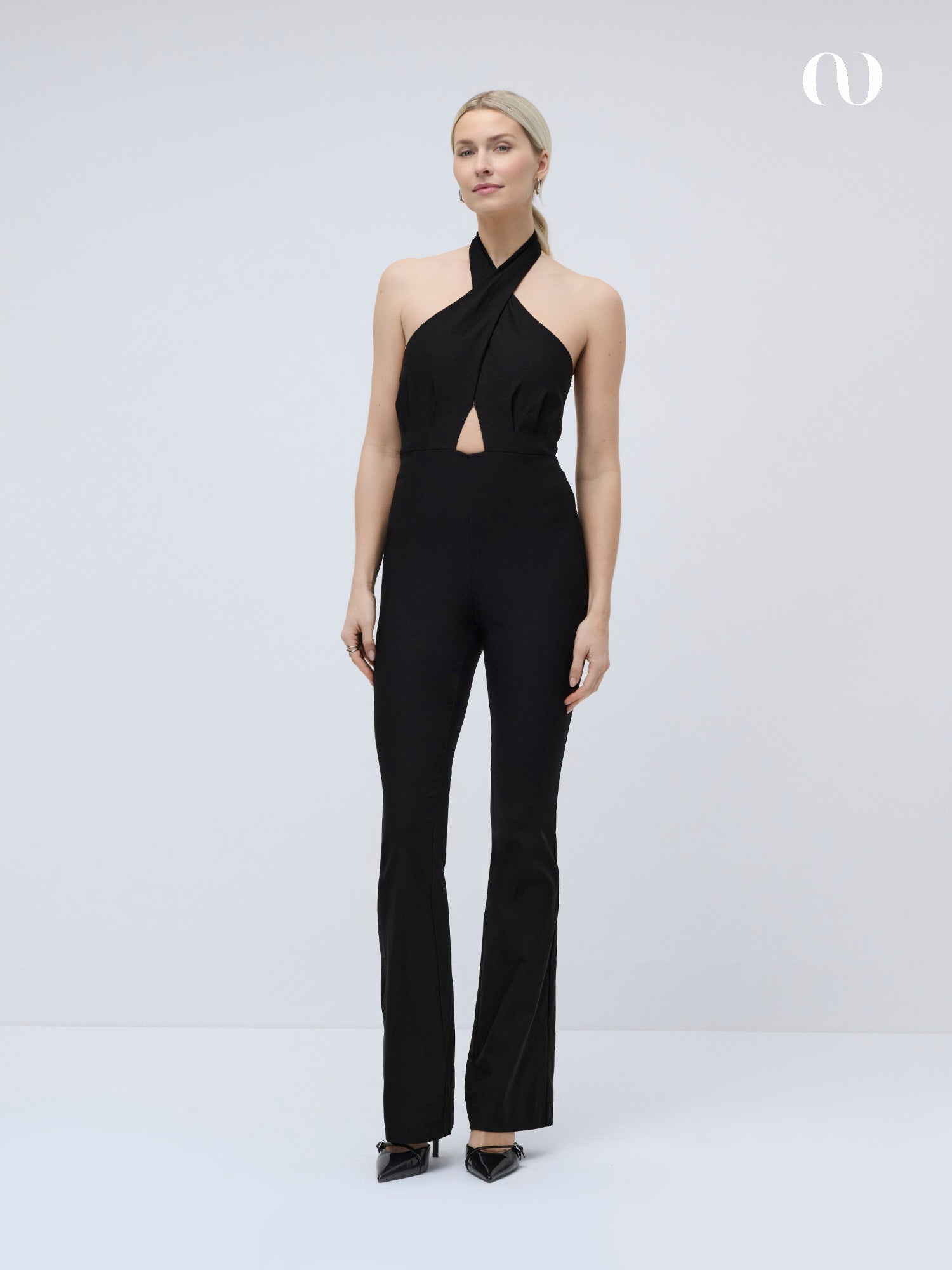 Eine Frau posiert vor einem schlichten, hellen Hintergrund. Sie trägt den LeGer by Lena Gercke Hacer Overall - einen schmal geschnittenen, schwarzen Trägeroverall mit einem Schlüssellochausschnitt auf der Brust - und dazu schwarze, spitze Absätze.