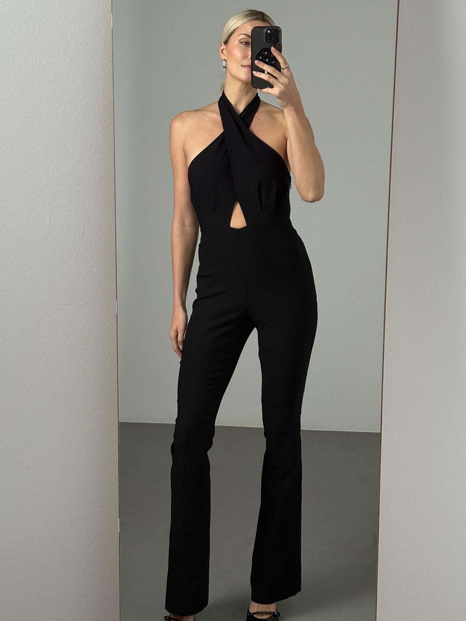 Eine Frau macht ein Spiegel-Selfie in einem LeGer by Lena Gercke Hacer Overall mit schmaler Passform, Neckholder und Taillenausschnitt in einem minimalistischen Raum mit schlichten Wänden und Boden.