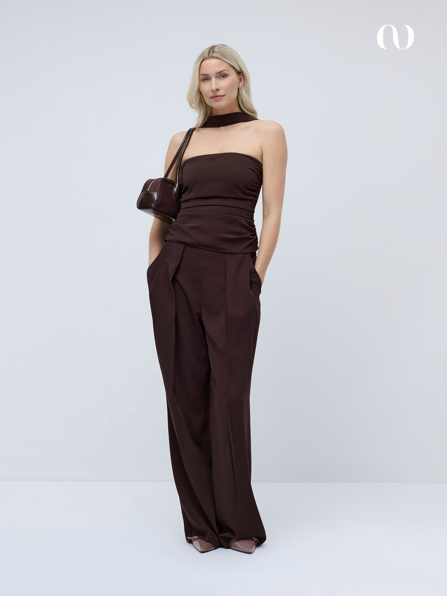 Eine Frau steht vor einem schlichten, hellen Hintergrund und trägt das Christina Top von LeGer by Lena Gercke, kombiniert mit einer passenden Hose mit weitem Bein, einem Halsband, spitzen Absätzen und einer kleinen Umhängetasche.