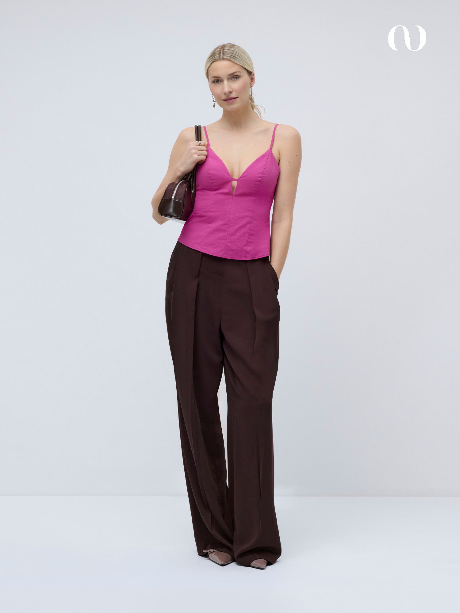 Eine Frau posiert vor einem schlichten Hintergrund in dem rosafarbenen Jella Top von LeGer by Lena Gercke mit Ausschnitt und tiefem V-Ausschnitt, kombiniert mit einer braunen Hose mit weitem Bein, einer passenden Handtasche, hochgestecktem Haar und Ohrringen.
