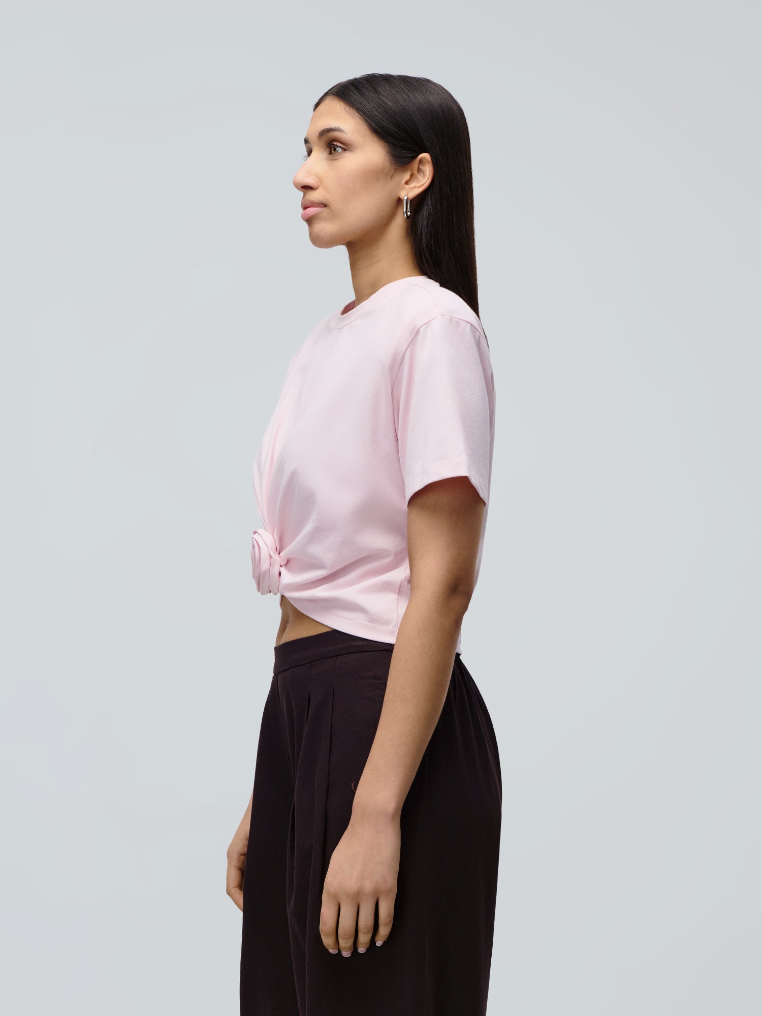 Eine Frau steht im Profil vor einem hellgrauen Hintergrund. Sie trägt das „Ina“-T-Shirt von LeGer by Lena Gercke in Hellrosa, das vorne zusammengebunden ist, dazu eine schwarze Hose. Ihr dunkles, glattes Haar fällt ihr offen über die Schultern, während sie nach vorne blickt.