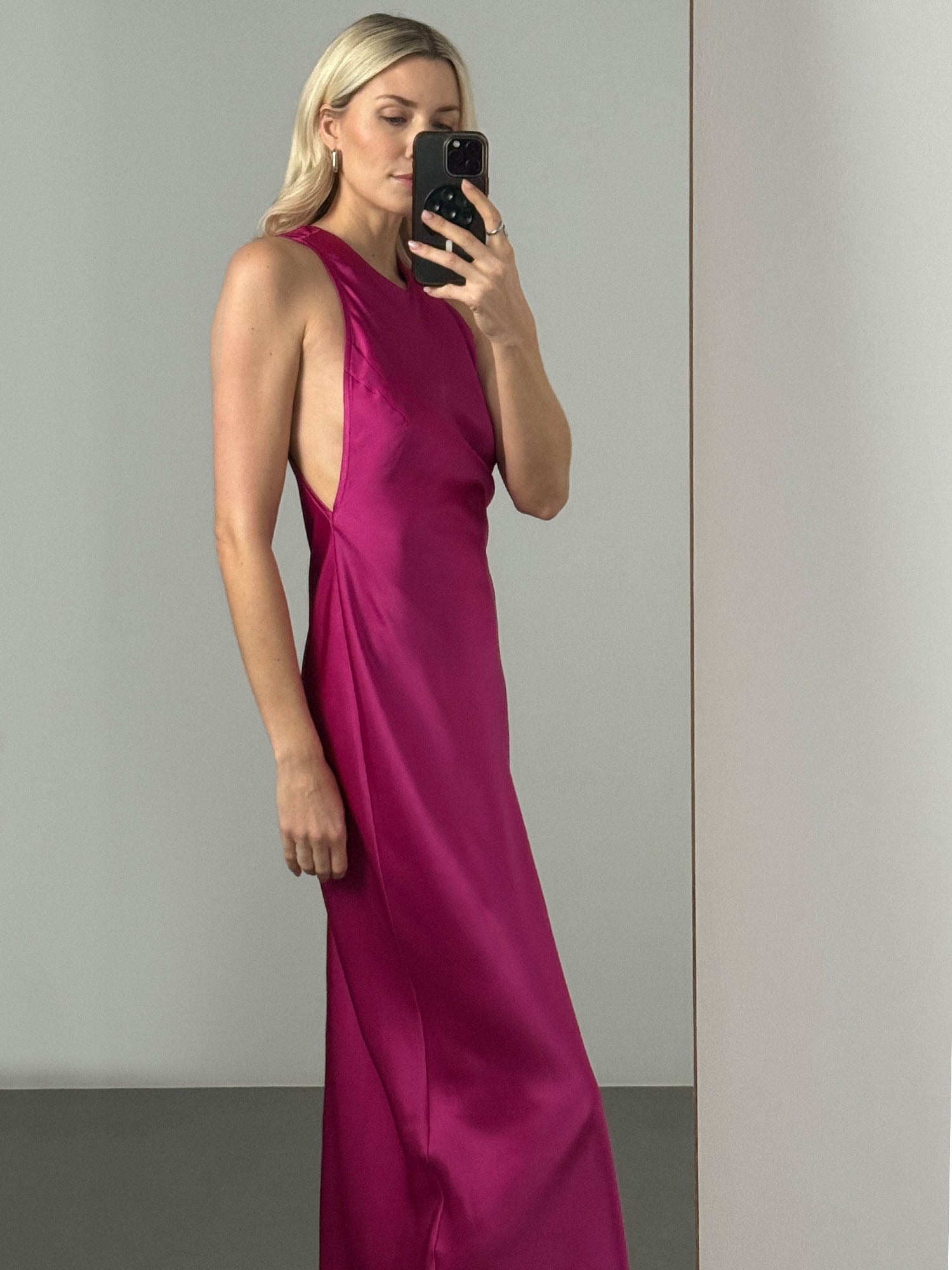 Eine Frau mit langen blonden Haaren macht ein Spiegel-Selfie in einem minimalistischen, neutral getönten Raum. Sie trägt das fuchsiafarbene Else Dress von LeGer by Lena Gercke - ein ärmelloses Maxikleid aus Satin mit tiefem Seitenausschnitt.