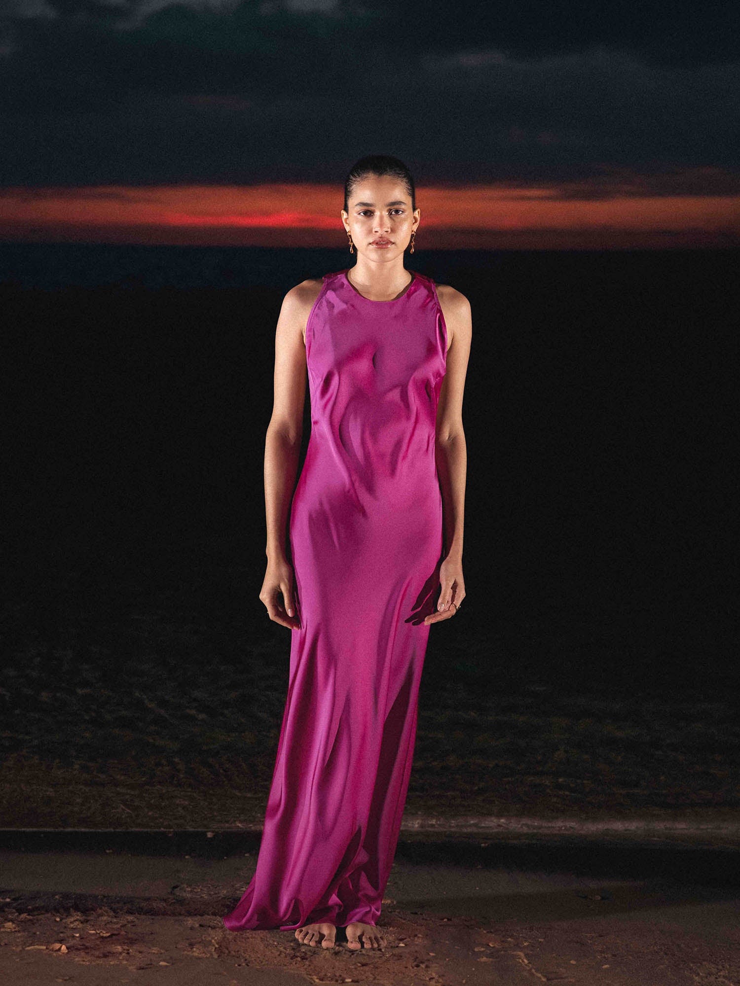 Eine Frau steht bei Sonnenuntergang barfuß am Ufer und trägt das leuchtend magentafarbene Else Dress von LeGer by Lena Gercke mit Rundhalsausschnitt. Der dunkle Himmel leuchtet in der Nähe des Horizonts rot, während das Wasser hinter ihr schimmert.