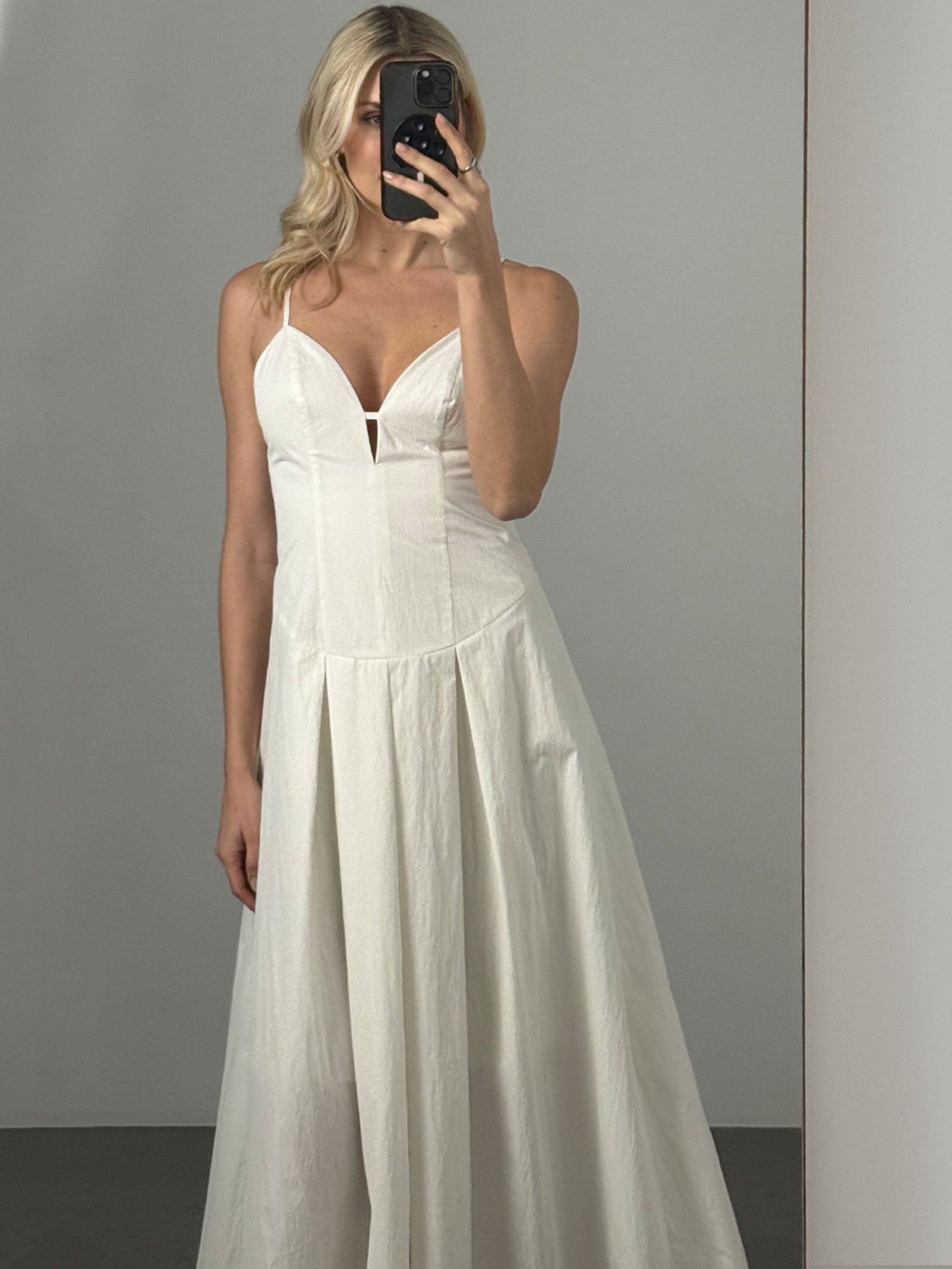 Eine Frau mit langen blonden Haaren macht ein Selfie im Spiegel. Sie trägt das knöchellange Jella Dress von LeGer by Lena Gercke, ein ärmelloses, fließendes weißes Kleid mit Kellerfalten, einem taillierten Mieder und einem dezenten V-Ausschnitt.