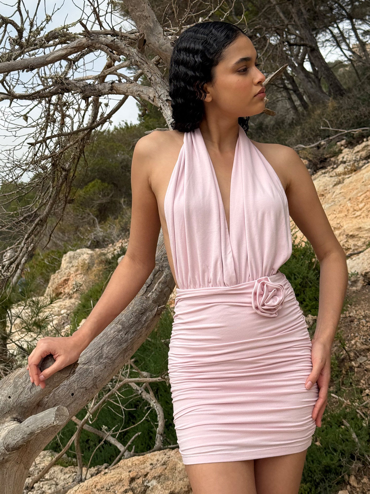 Eine Frau steht im Freien in der Nähe von Felsen und Bäumen und trägt das Kleid „Rosie“ von LeGer by Lena Gercke – ein hellrosa, ärmelloses Neckholder-Kleid mit gerafftem Rock und einer Stoffblume an der Taille – und blickt nach rechts.