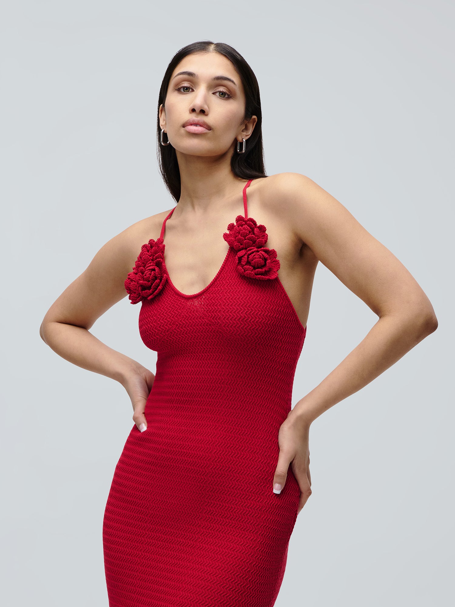 Eine Frau posiert selbstbewusst in dem ärmellosen roten Strickkleid „Lia“ von LeGer by Lena Gercke, das sich durch eine Meerjungfrauen-Silhouette und florale Verzierungen im Brustbereich auszeichnet, vor einem schlichten hellgrauen Hintergrund.