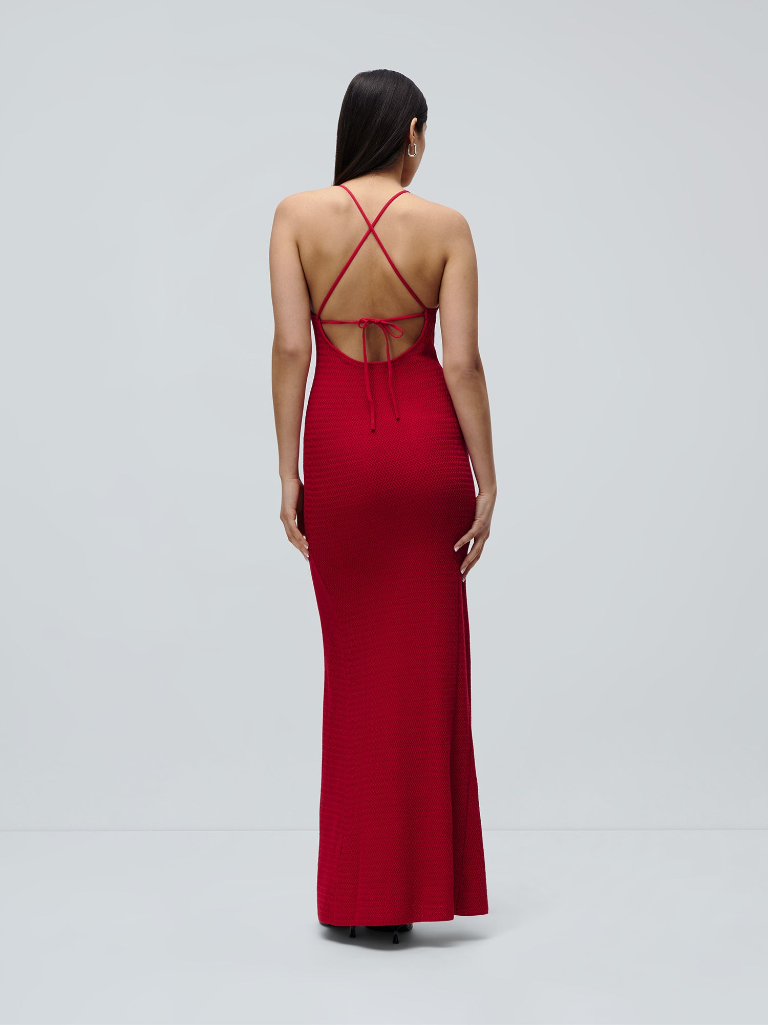 Eine Frau mit langen, dunklen Haaren ist von hinten zu sehen. Sie trägt das ärmellose, bodenlange „Lia“-Kleid von LeGer by Lena Gercke mit offenem Rücken und gekreuzten Trägern. Der Hintergrund ist einfarbig hellgrau.
