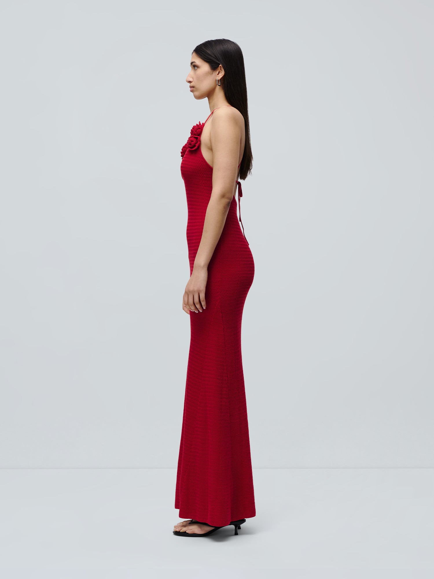 Eine Frau posiert im Profil vor einem hellgrauen Hintergrund. Sie trägt das lange, figurbetonte Kleid „Lia“ von LeGer by Lena Gercke mit floralen Verzierungen im Brustbereich sowie schwarze Sandalen mit Absatz.