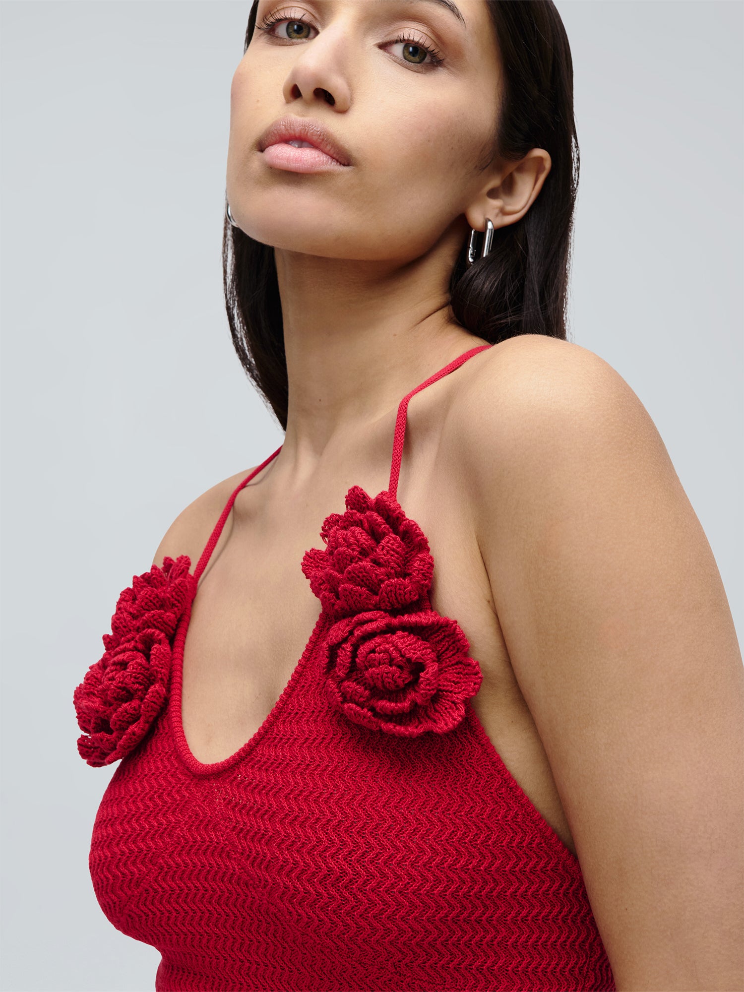 Eine Frau mit langen, dunklen Haaren trägt das Kleid „Lia“ von LeGer by Lena Gercke, ein rotes, strukturiertes Kleid mit schmalen Trägern und großen, gehäkelten Blumenverzierungen. Vor einem schlichten, hellen Hintergrund blickt sie mit neutralem Gesichtsausdruck leicht nach oben.