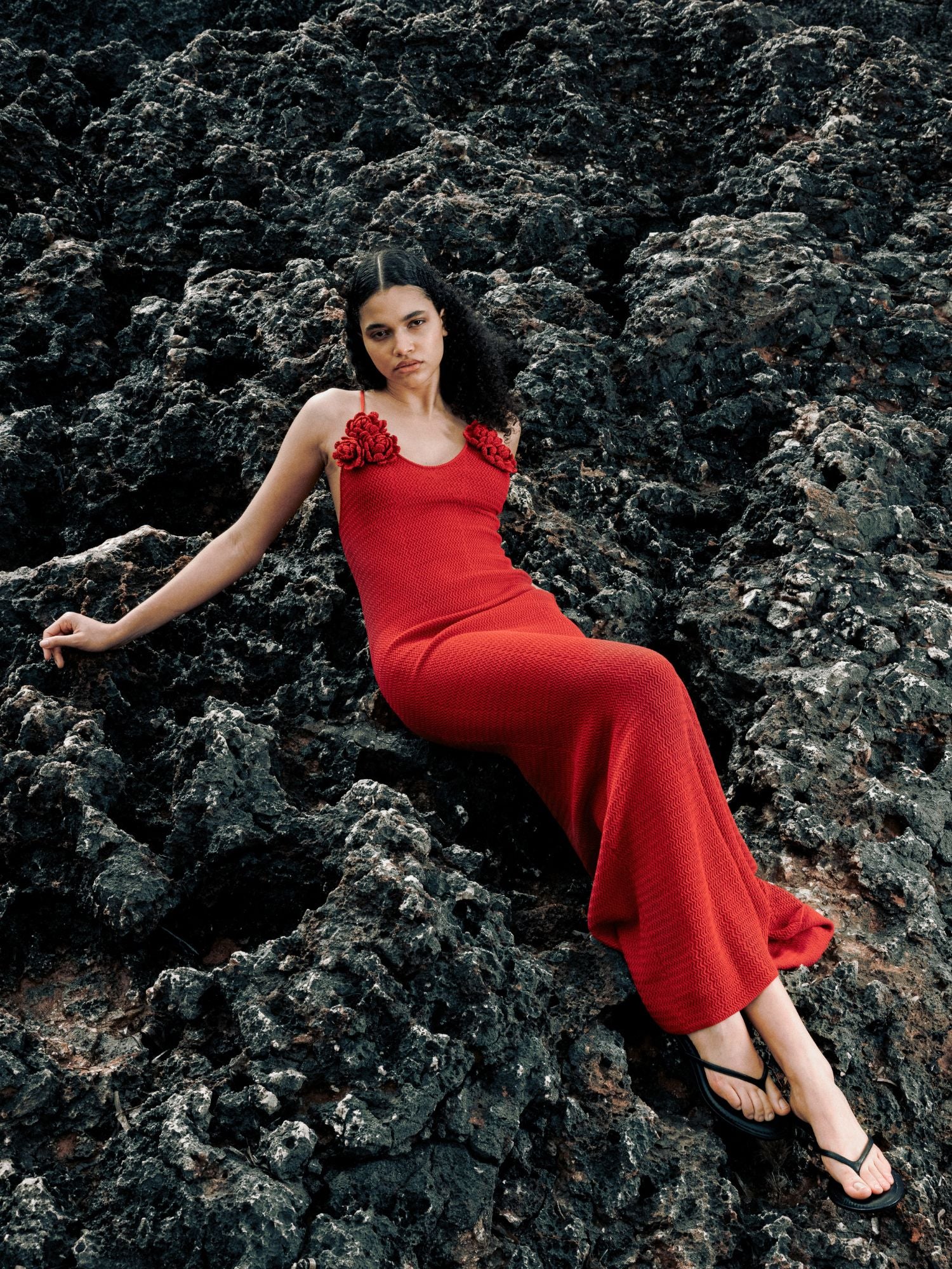 Eine Frau trägt das rote "Lia Dress" von LeGer by Lena Gercke mit Rosen-Details an den Trägern, liegt entspannt auf dunklem Vulkangestein, trägt schwarze Sandalen und blickt ruhig in die Kamera.
