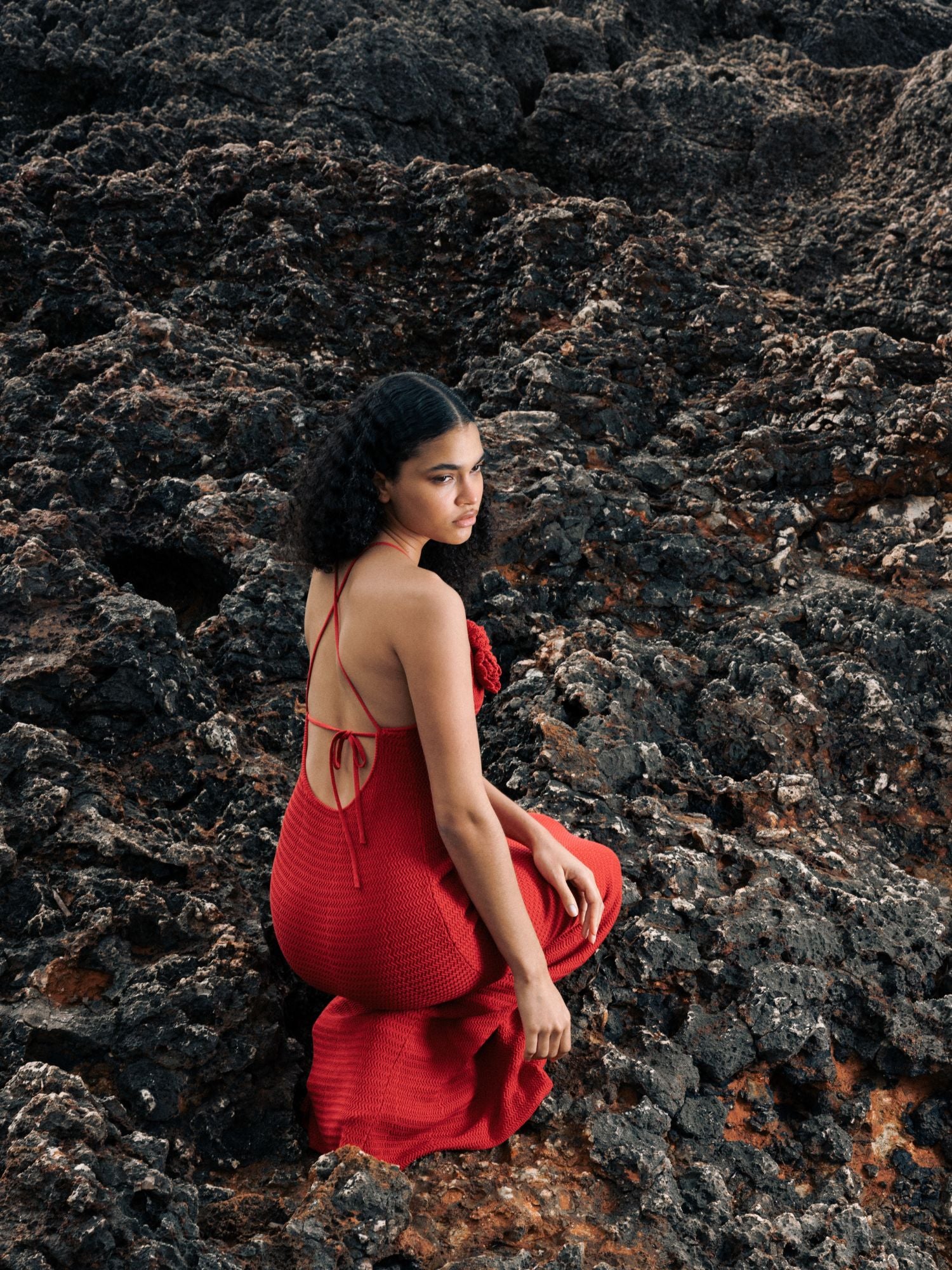 Eine Frau, die das Kleid „Lia“ von LeGer by Lena Gercke trägt, kniet auf dunklen Vulkangesteinen, ihren ernsten Blick zur Seite gerichtet.