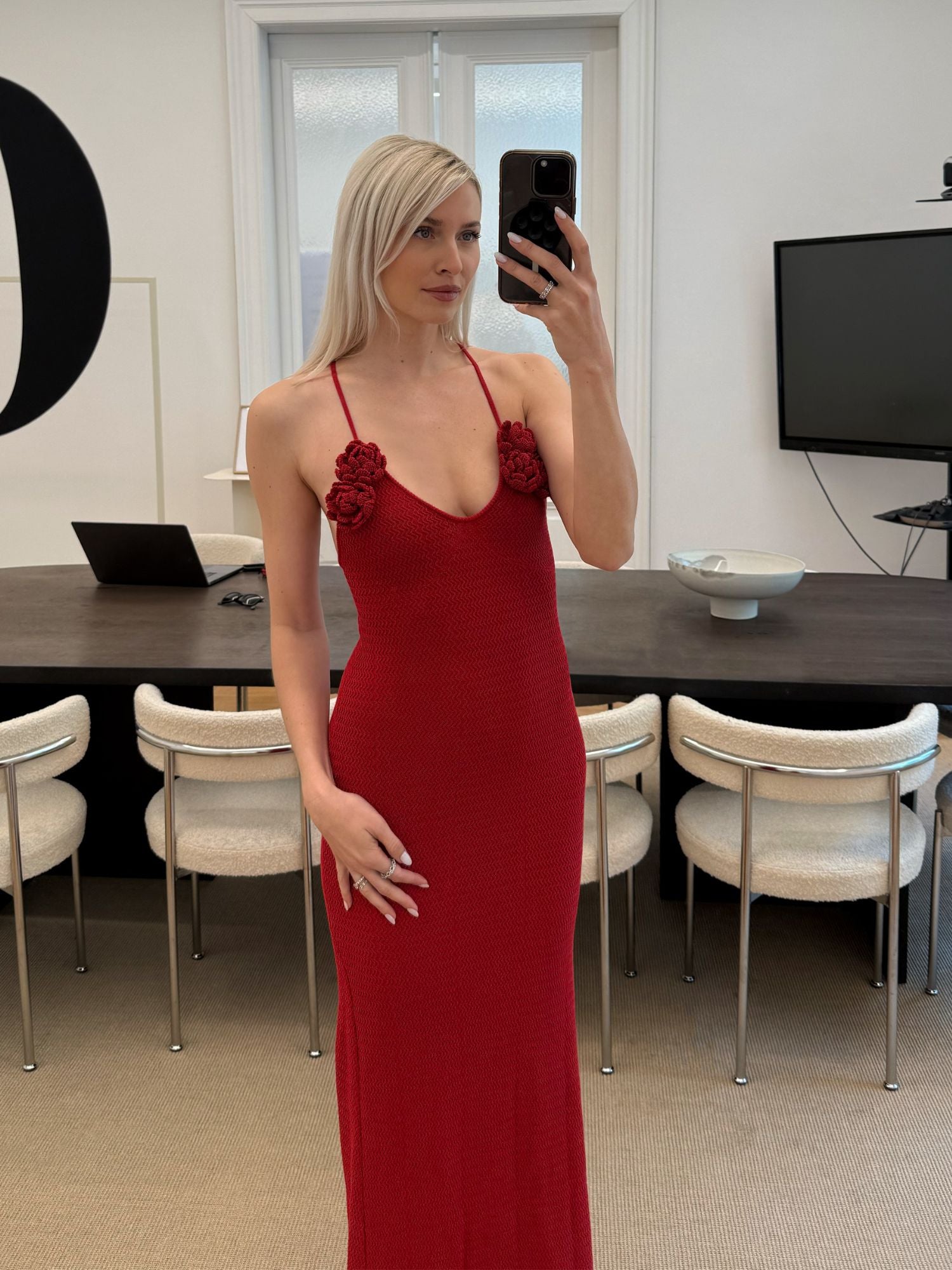 Eine Frau mit langen blonden Haaren macht in einem modernen Büro ein Selfie im Spiegel. Sie trägt das Lia Dress von LeGer by Lena Gercke - ein elegantes, ärmelloses, bodenlanges rotes Kleid mit geblümten Trägerakzenten. Hinter ihr stehen ein dunkler Tisch, weiße Stühle und ein großer Bildschirm.