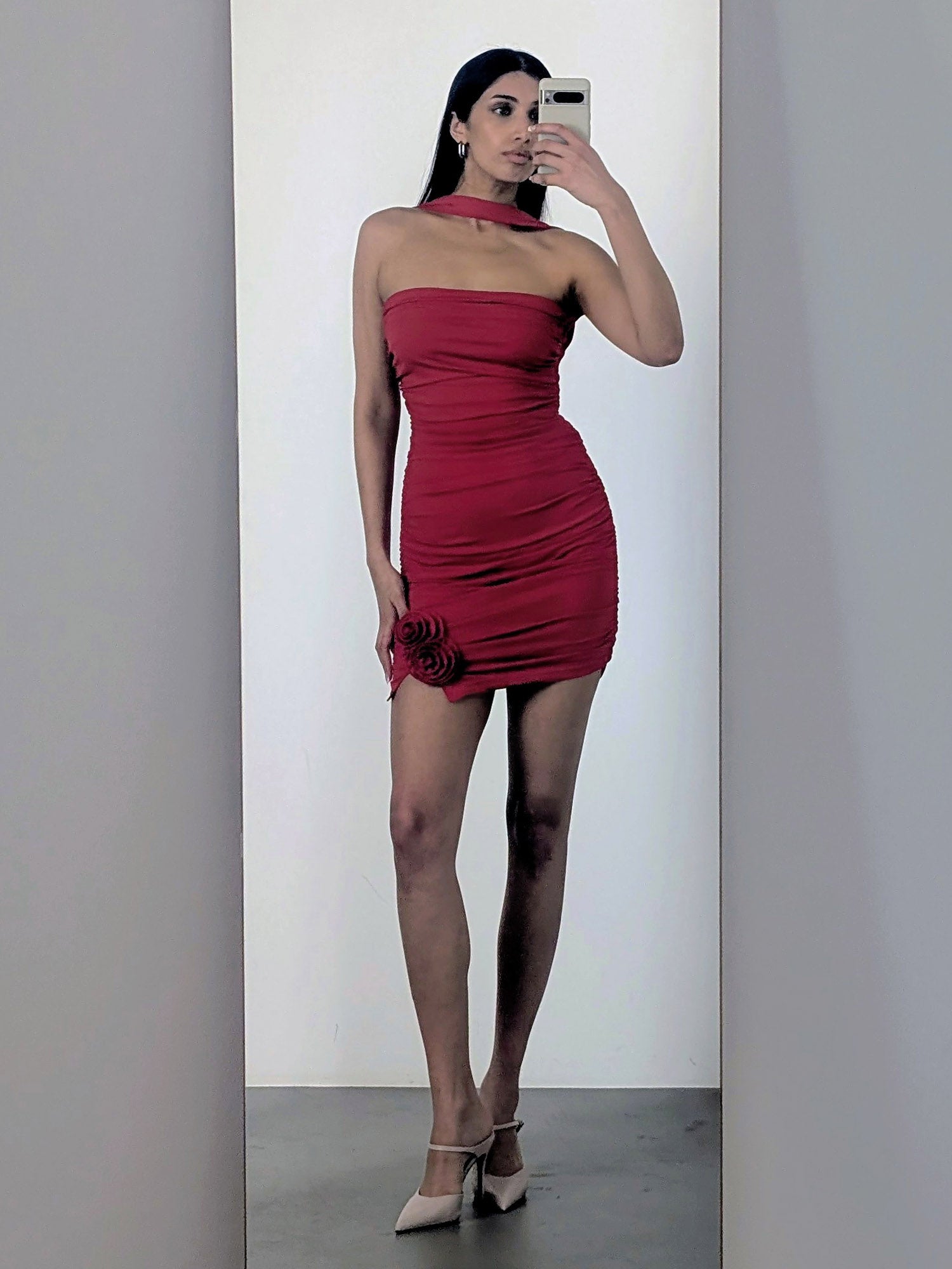 Eine Person steht vor einem Spiegel und macht ein Selfie. Sie trägt das Kleid „Eike“ von LeGer by Lena Gercke. Das eng anliegende, rote Minikleid mit Rosenverzierungen am Saum wird mit beigen Pumps kombiniert, wodurch sich ihre Silhouette vor einem minimalistischen Hintergrund besonders gut abhebt.