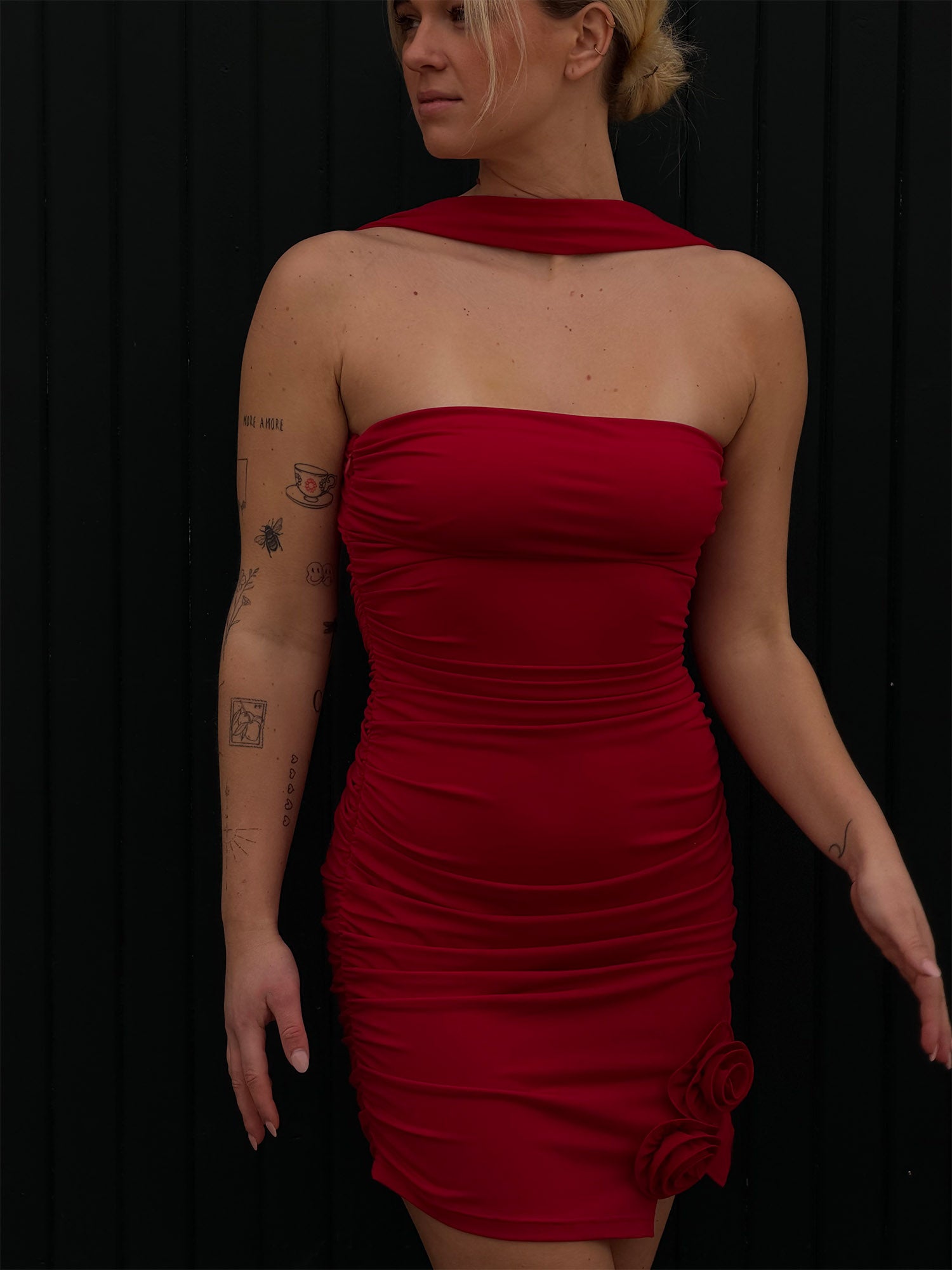 Eine Frau steht vor einer schwarzen Wand und trägt das Kleid „Eike“ von LeGer by Lena Gercke – ein trägerloses, figurbetontes rotes Minikleid mit gerafften Seiten und zwei Stoffrosen am Saum. Ihr blondes Haar ist nach hinten gebunden, sodass ihre Arm-Tattoos zu sehen sind.