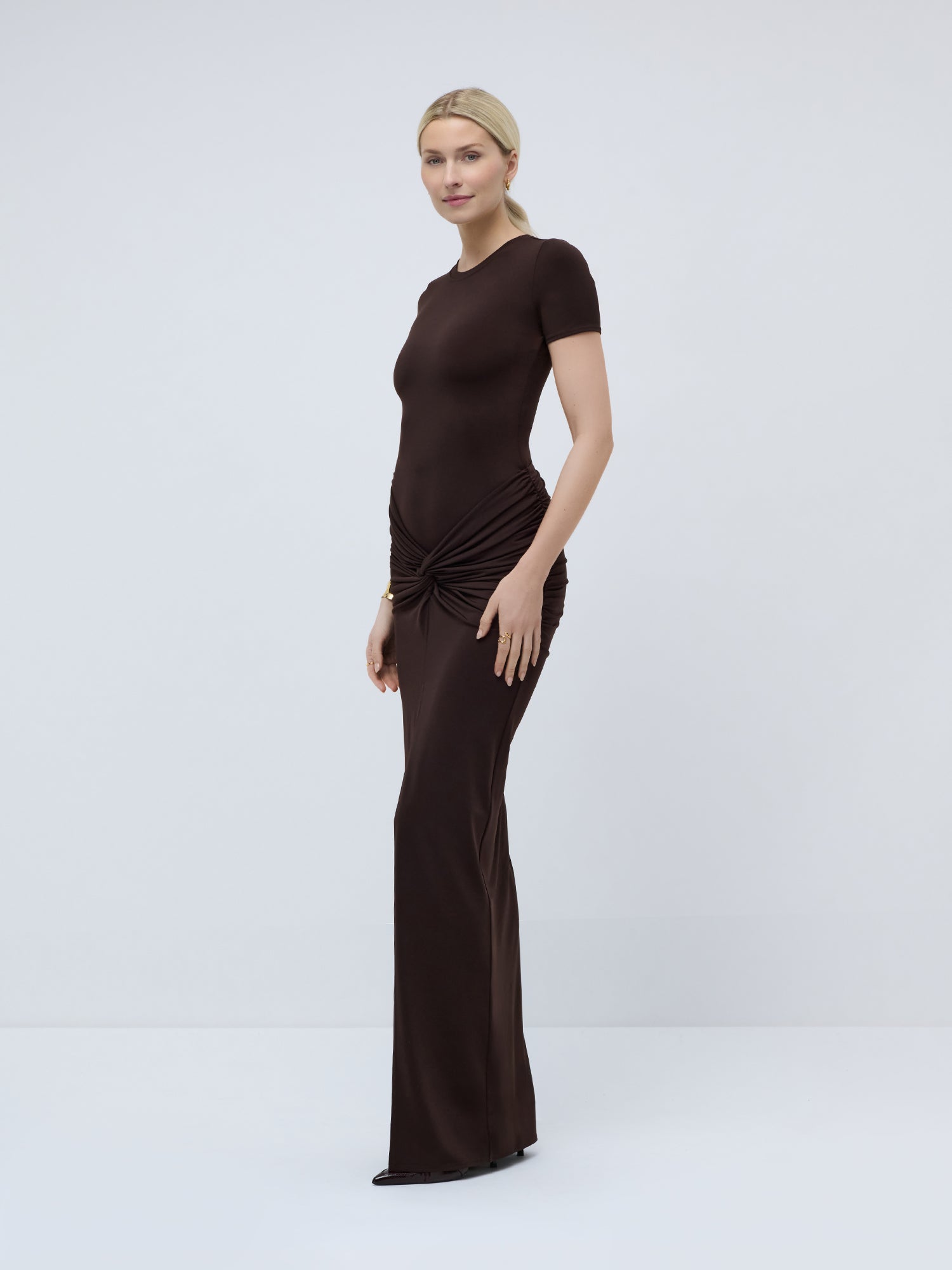 Eine Frau mit blonden Haaren, die zu einem tief sitzenden Pferdeschwanz zusammengebunden sind, trägt das figurbetonte, kurzärmelige, bodenlange braune „Gisa“-Kleid von LeGer by Lena Gercke mit Knotendetails an den Hüften, während sie vor einem schlichten hellgrauen Hintergrund steht.