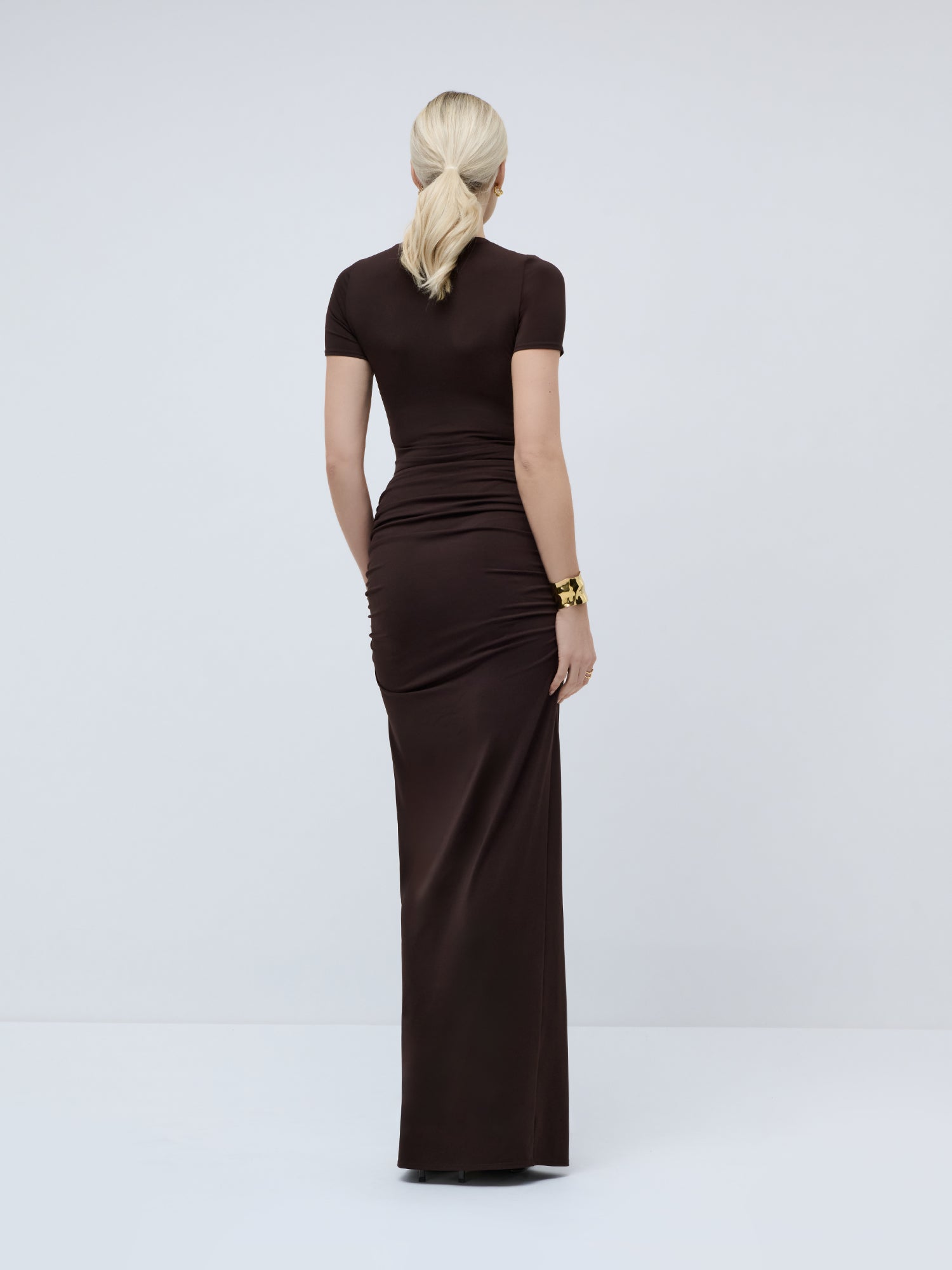 Eine blonde Frau mit einem tief gebundenen Pferdeschwanz steht mit dem Rücken zur Kamera und trägt das eng anliegende, dunkelbraune Maxikleid „Gisa“ mit kurzen Ärmeln und Knotendetail von LeGer by Lena Gercke sowie ein goldenes Armband, vor einem schlichten, hellen Hintergrund.