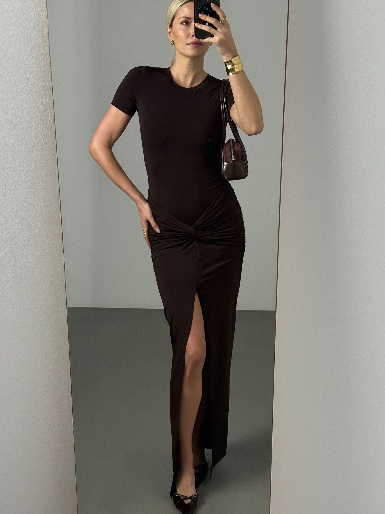 Eine Frau trägt das dunkelbraune „Gisa“-Kleid von LeGer by Lena Gercke mit kurzen Ärmeln, einem Knotendetail und einem hohen Schlitz. Sie rundet den Look mit einem goldenen Armreif und einer burgunderroten Umhängetasche ab.