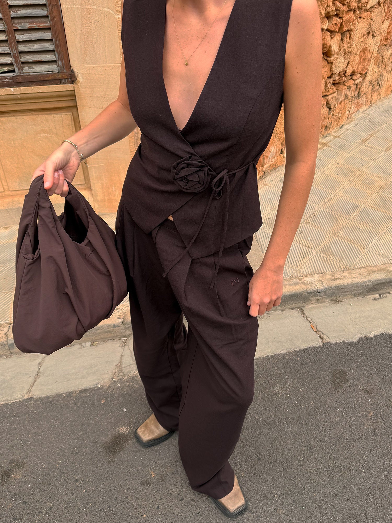 Eine Person in einem ärmellosen, dunkelbraunen Wickeloberteil mit Blumendetails, einer Jemie-Hose von LeGer by Lena Gercke und quadratischen Schuhen steht mit einer braunen Tasche in der Hand auf einem Gehweg in der Nähe einer hellbraunen Steinwand und eines Fensters.