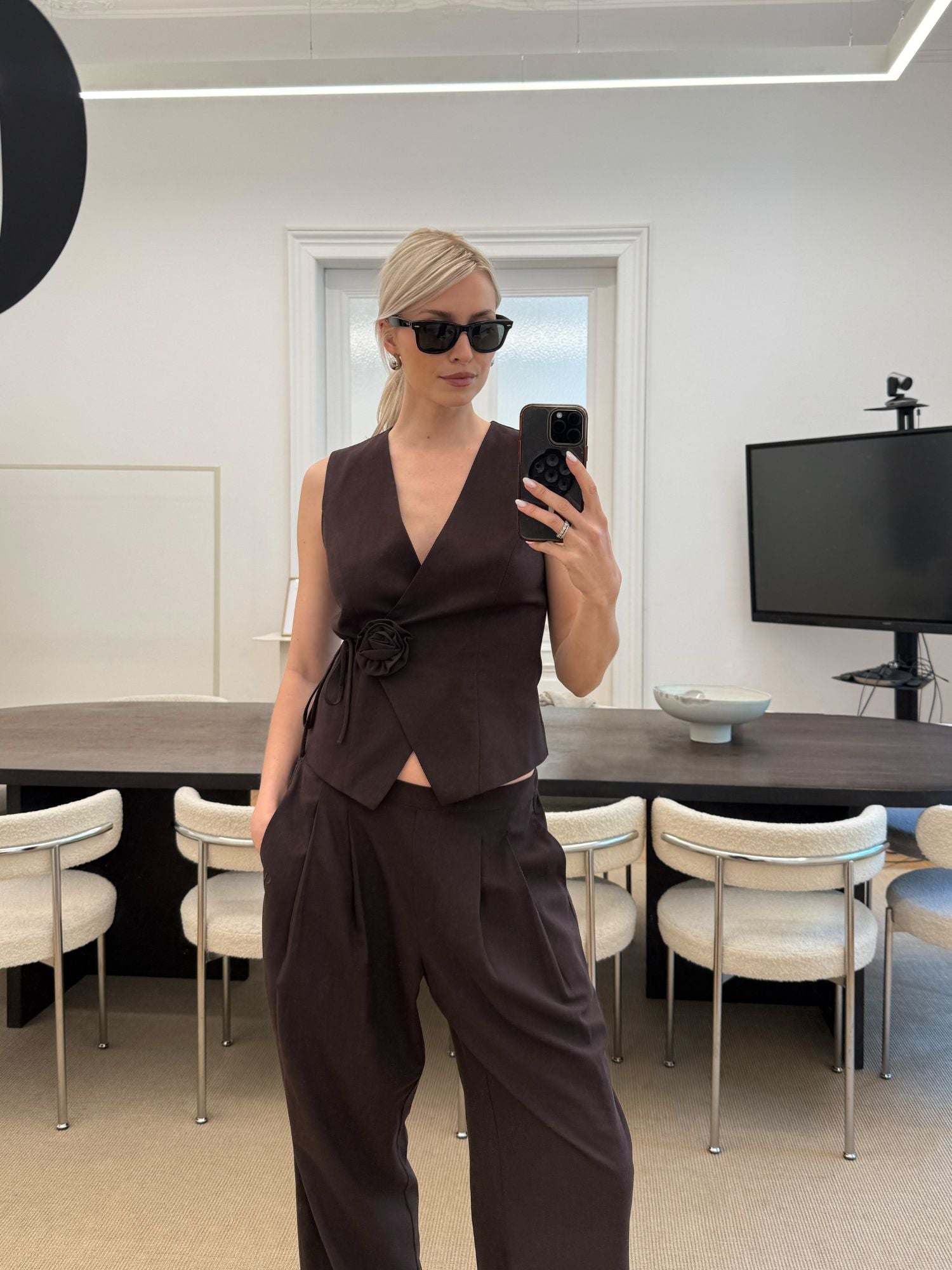 Eine Frau mit dunkler Sonnenbrille, ärmelloser brauner Weste und passender LeGer by Lena Gercke Jemie Pants macht ein Spiegel-Selfie in einem modernen Büro mit weißen Stühlen und einem großen wandmontierten Fernseher.