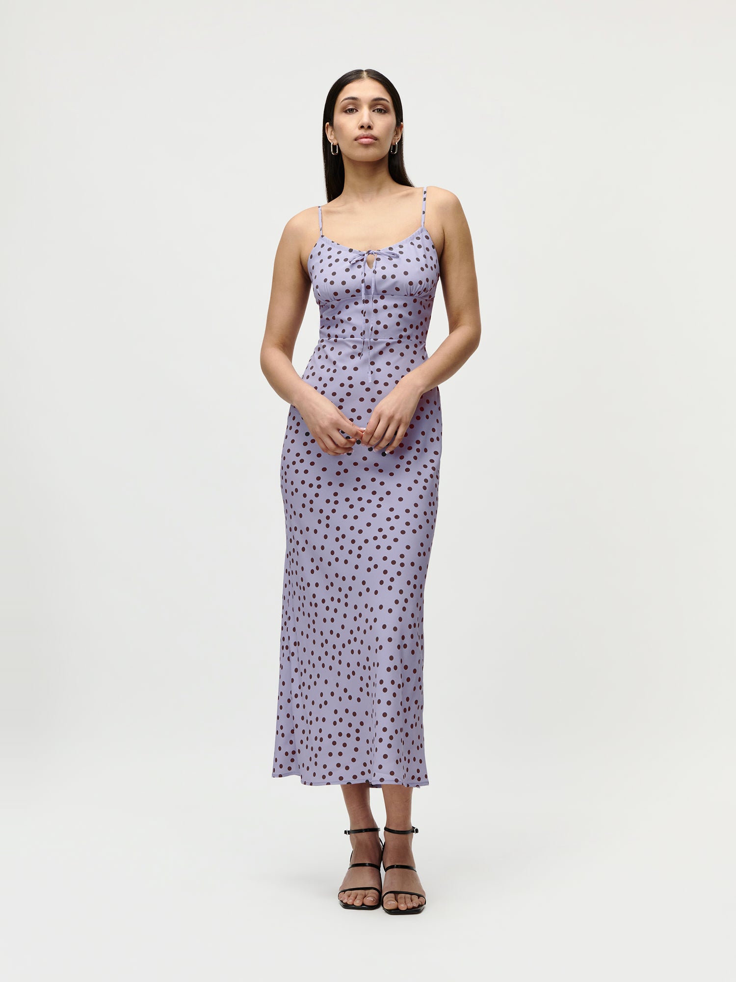 Eine Frau blickt nach vorne in dem hellvioletten, ärmellosen Fleur-Kleid mit schwarzen Tupfen und dünnen Trägern von LeGer by Lena Gercke, gestylt mit schwarzen Riemchensandalen - ein schicker Sommerlook vor einem schlichten weißen Hintergrund.