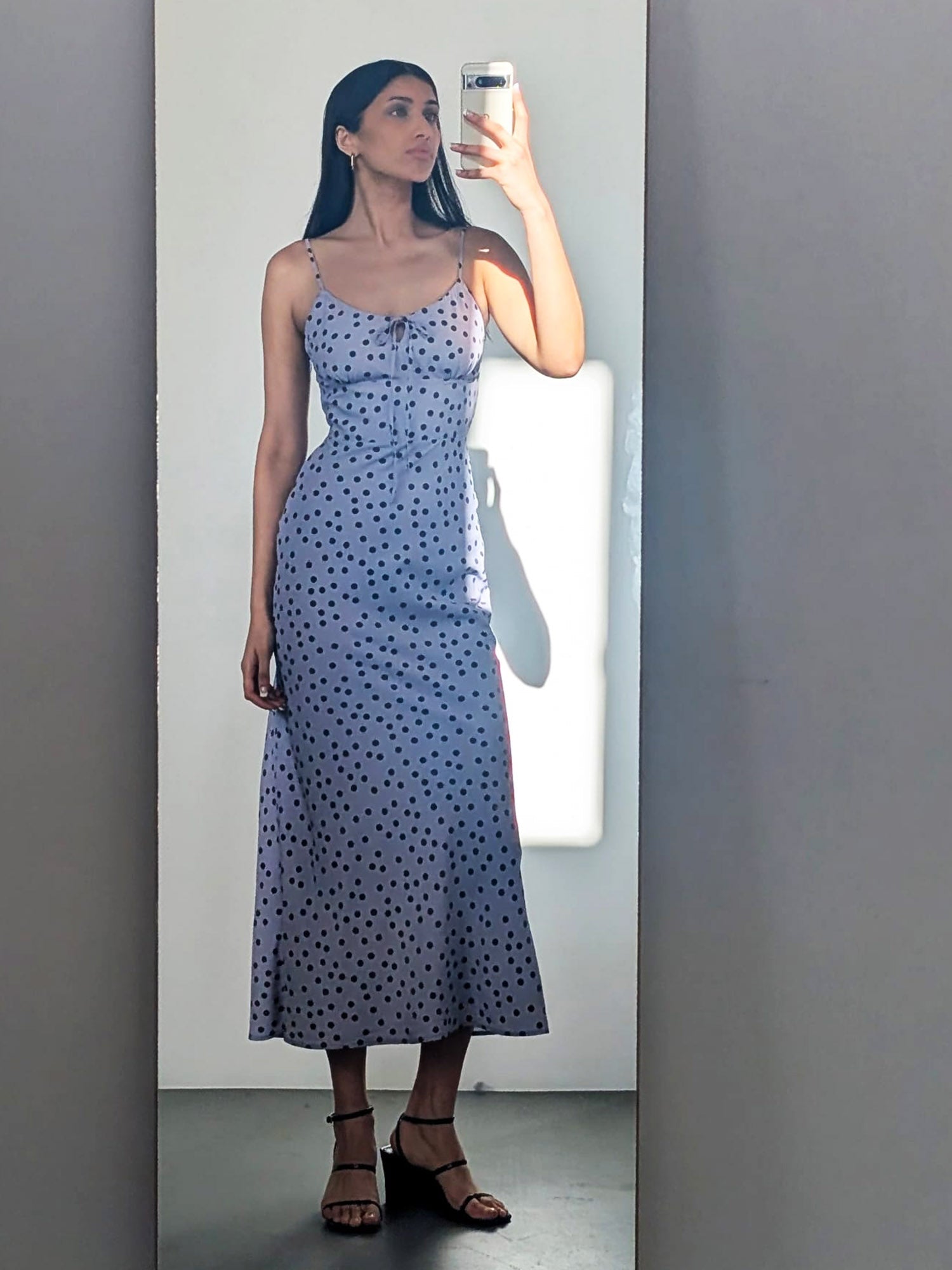 Eine Frau macht ein Selfie im Fleur Dress von LeGer by Lena Gercke - einem hellvioletten, knöchellangen Kleid mit schwarzen Tupfen - und vervollständigt ihren Look mit schwarzen Riemchensandalen und Sonnenlicht, das einen Schatten hinter sie wirft.