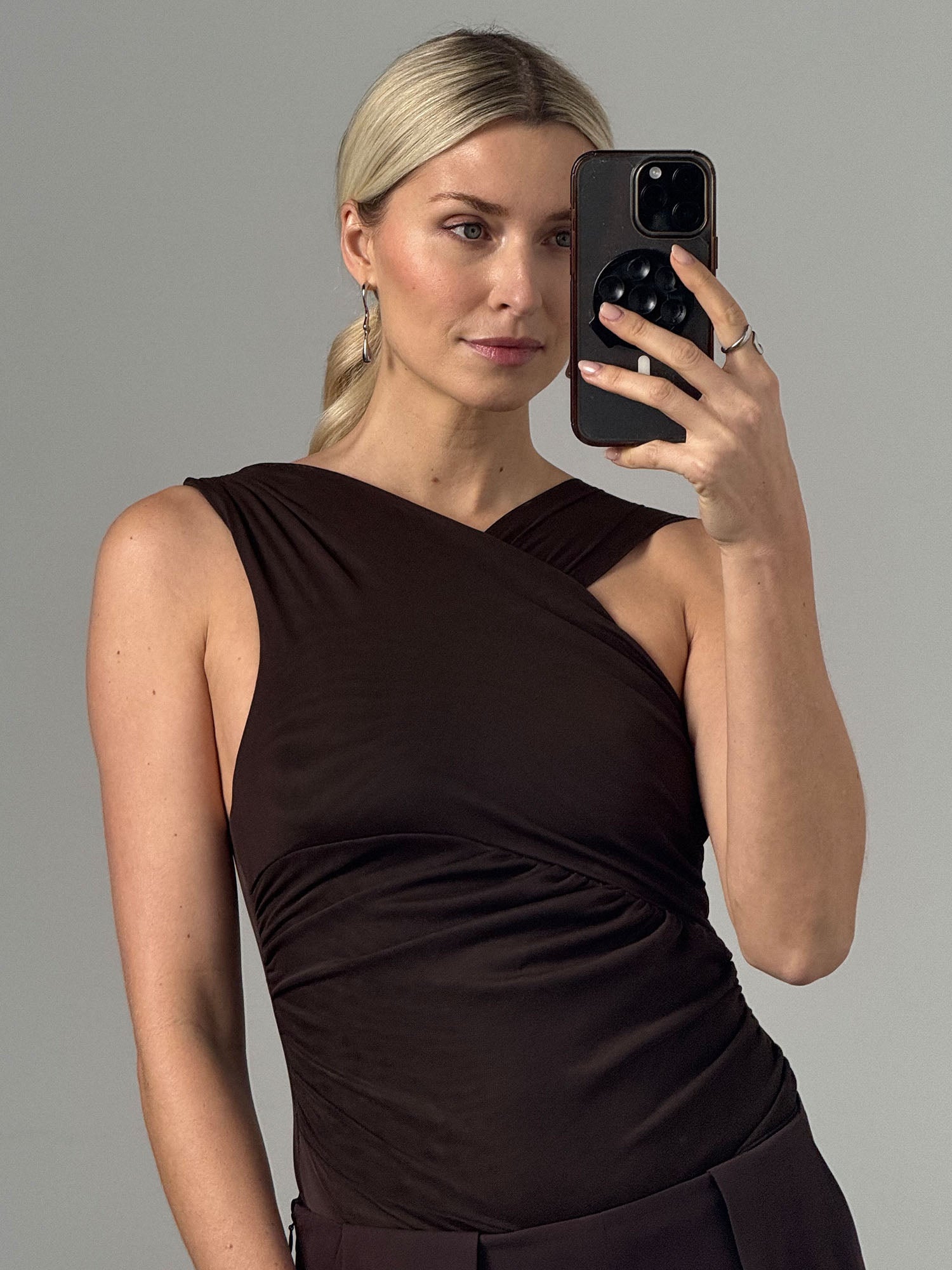 Eine Frau mit blonden Haaren im niedrigen Zopf macht ein Selfie vor grauem Hintergrund. Sie trägt das ärmellose, figurbetonte Dinorah Body von LeGer by Lena Gercke in Dunkelbraun (Größe 36), silberne Creolen und hält ein schwarzes Smartphone.
