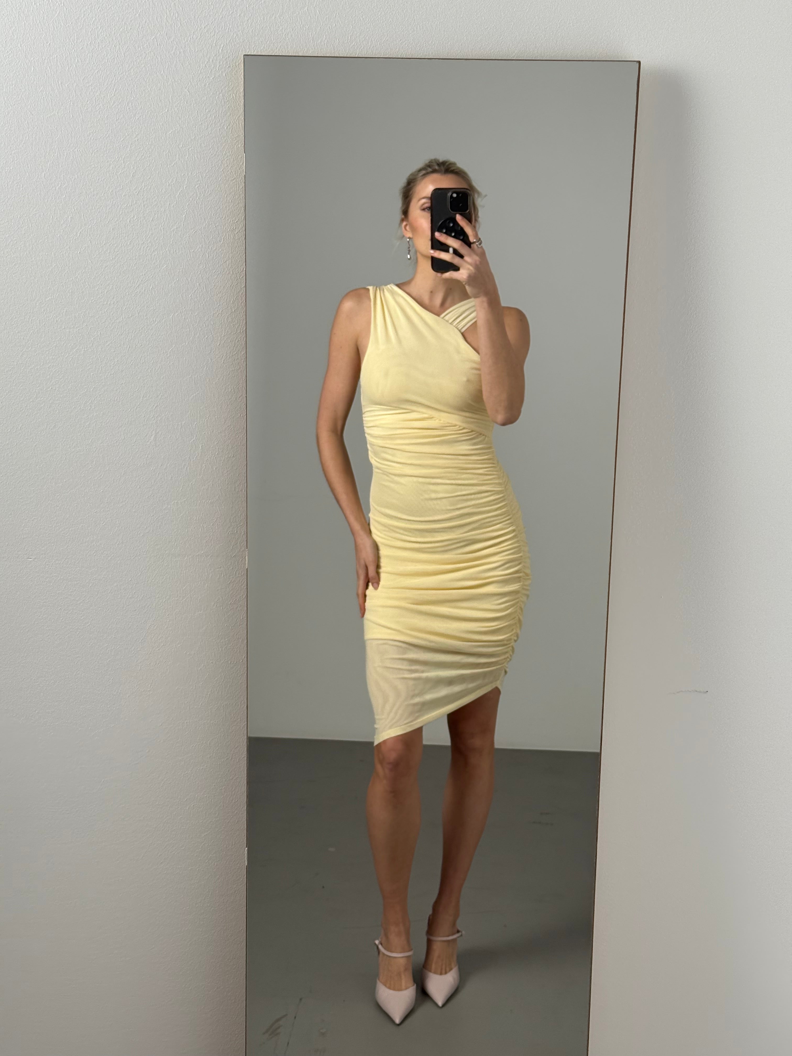 Eine Frau macht ein Selfie im Dinorah Dress von LeGer by Lena Gercke, einem blassgelben, ärmellosen, schulterfreien Midikleid mit Rüschen, das mit hellen Absätzen in einem minimalistischen Raum mit hellgrauem Boden und weißen Wänden kombiniert wird.
