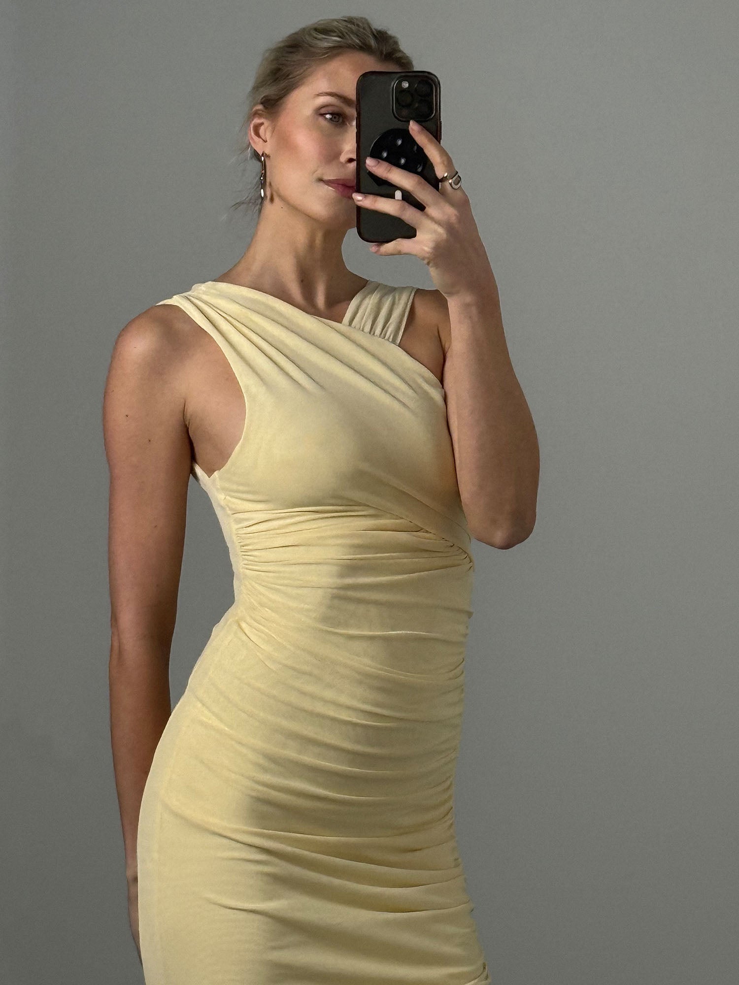 Eine Frau in dem Dinorah-Kleid von LeGer by Lena Gercke, einem blassgelben, ärmellosen, figurbetonten Midikleid mit einer Schulter, macht ein Selfie vor einem grauen Hintergrund. Ihr helles Haar ist zurückgesteckt und sie trägt Ohrringe.