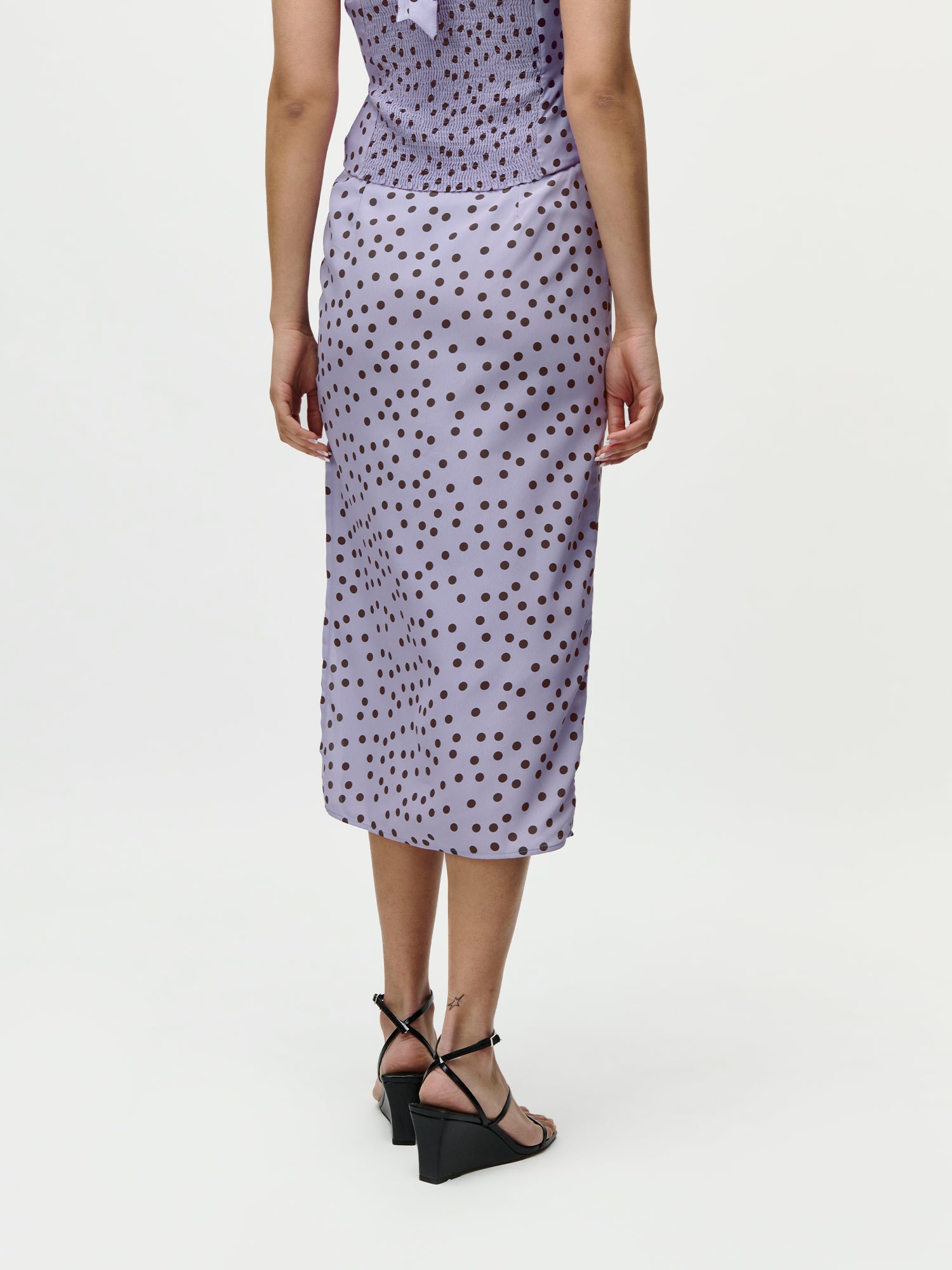 Eine Frau, die von der Seite schaut, trägt das LeGer by Lena Gercke Fleur Set - einen hellvioletten, gepunkteten Midirock und ein passendes Oberteil - das als Set in Größe 36 zu einem attraktiven Preis erhältlich ist. Der Hintergrund ist einfarbig weiß.