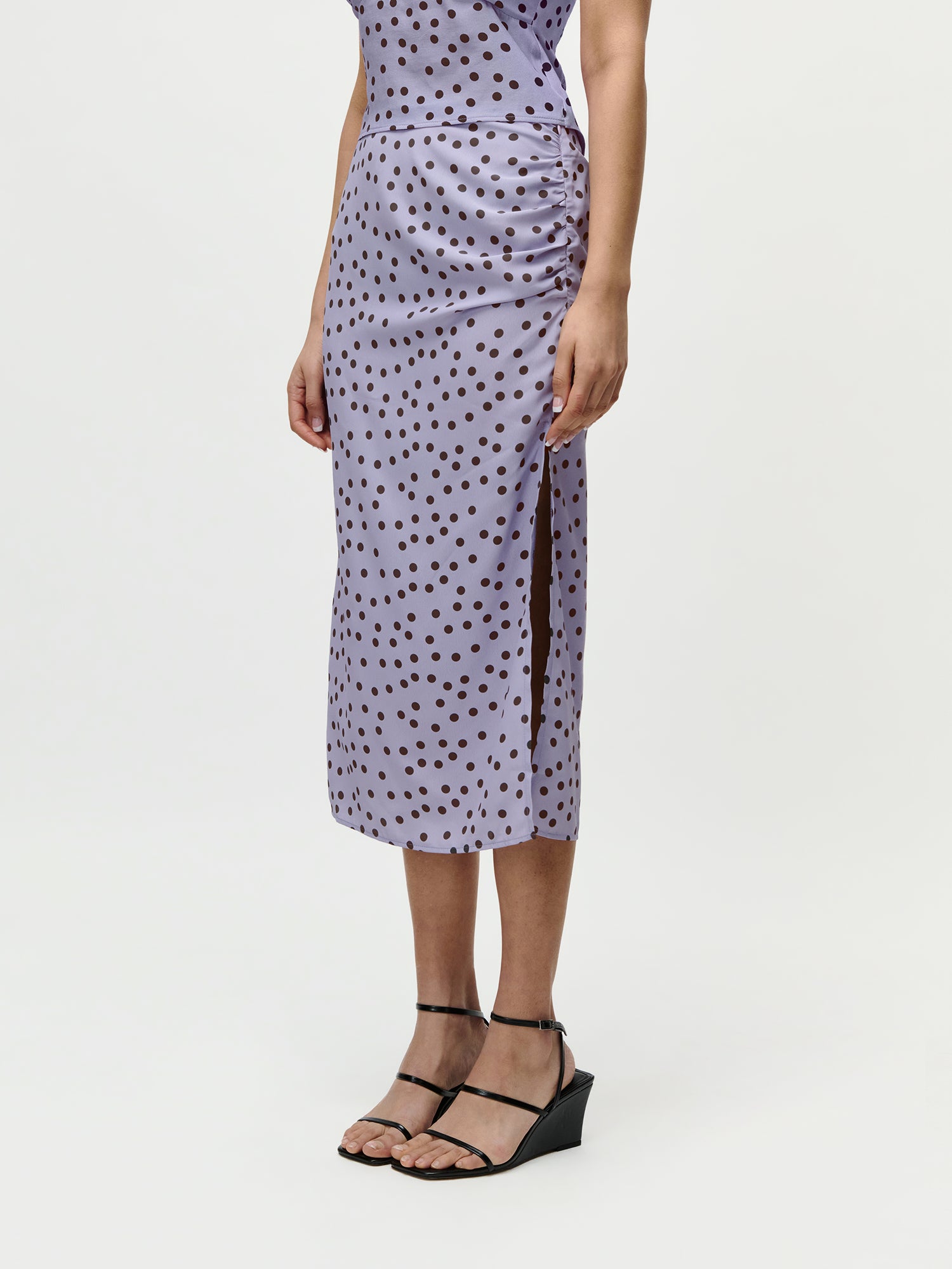 Eine Person modelliert das Fleur Set von LeGer by Lena Gercke - ein lavendelfarbener Midirock mit schwarzen Tupfen (Größe 36), seitlich geschlitzt - gepaart mit schwarzen Riemchen-Keilsandalen, vor einem schlichten weißen Hintergrund.