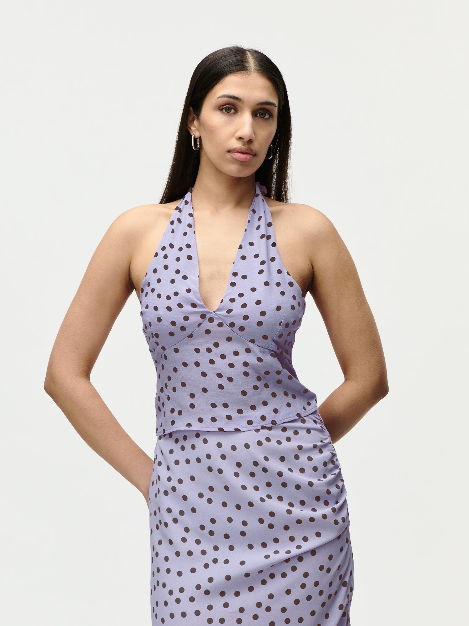 Eine Frau mit langen dunklen Haaren trägt das LeGer by Lena Gercke Fleur Set in Größe 36 - ein ärmelloses lavendelfarbenes Kleid mit Neckholder und schwarzen Punkten - vor einem schlichten hellen Hintergrund und blickt neutral in die Kamera.