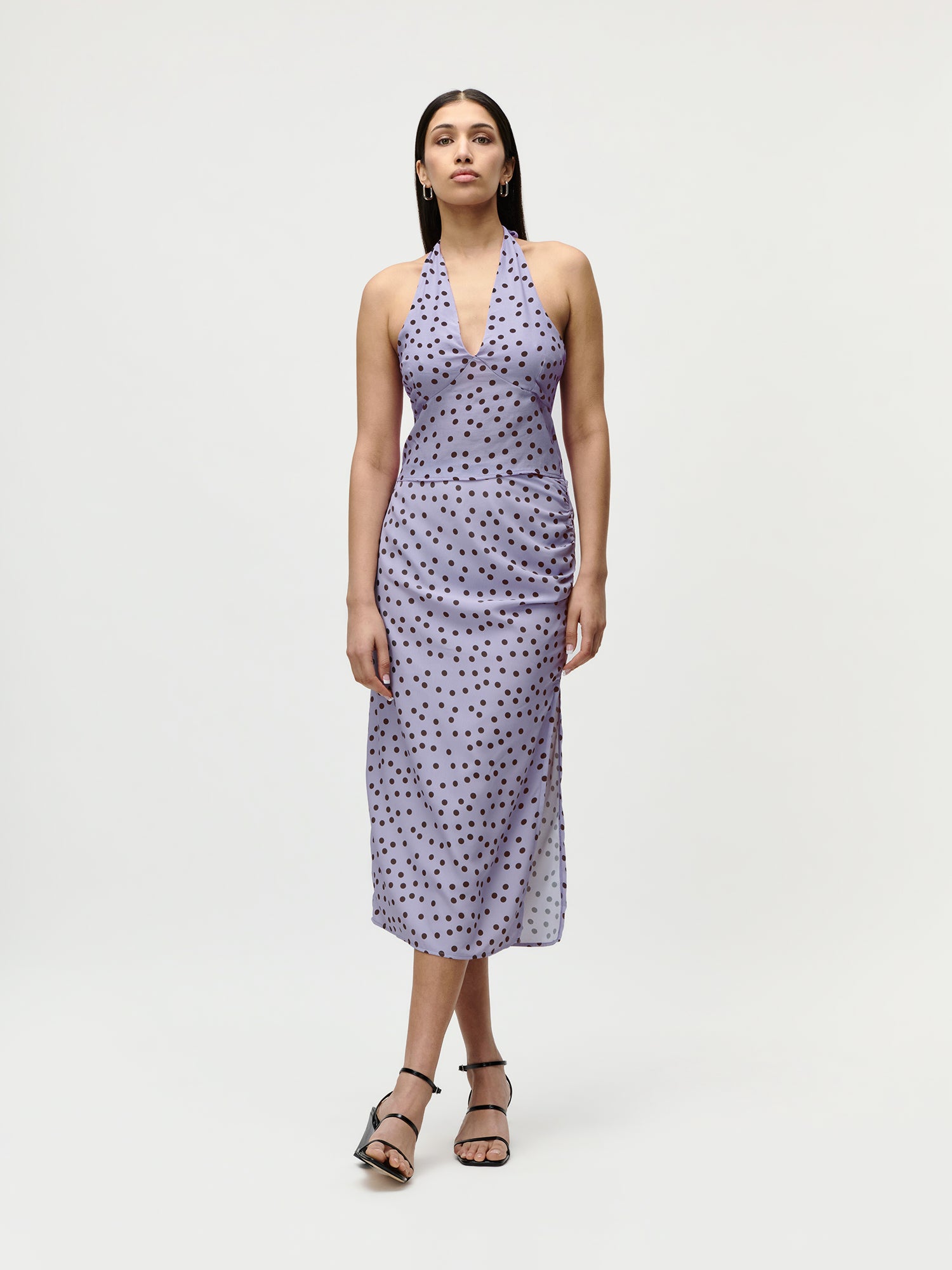 Eine Frau mit glattem dunklem Haar trägt das Fleur Set von LeGer by Lena Gercke: ein hellviolettes Midikleid mit Neckholder und schwarzen Tupfen (Größe 36) und schwarzen Riemchenabsätzen - ideal, wenn Sie das Set kaufen und Preisvorteile sichern möchten.