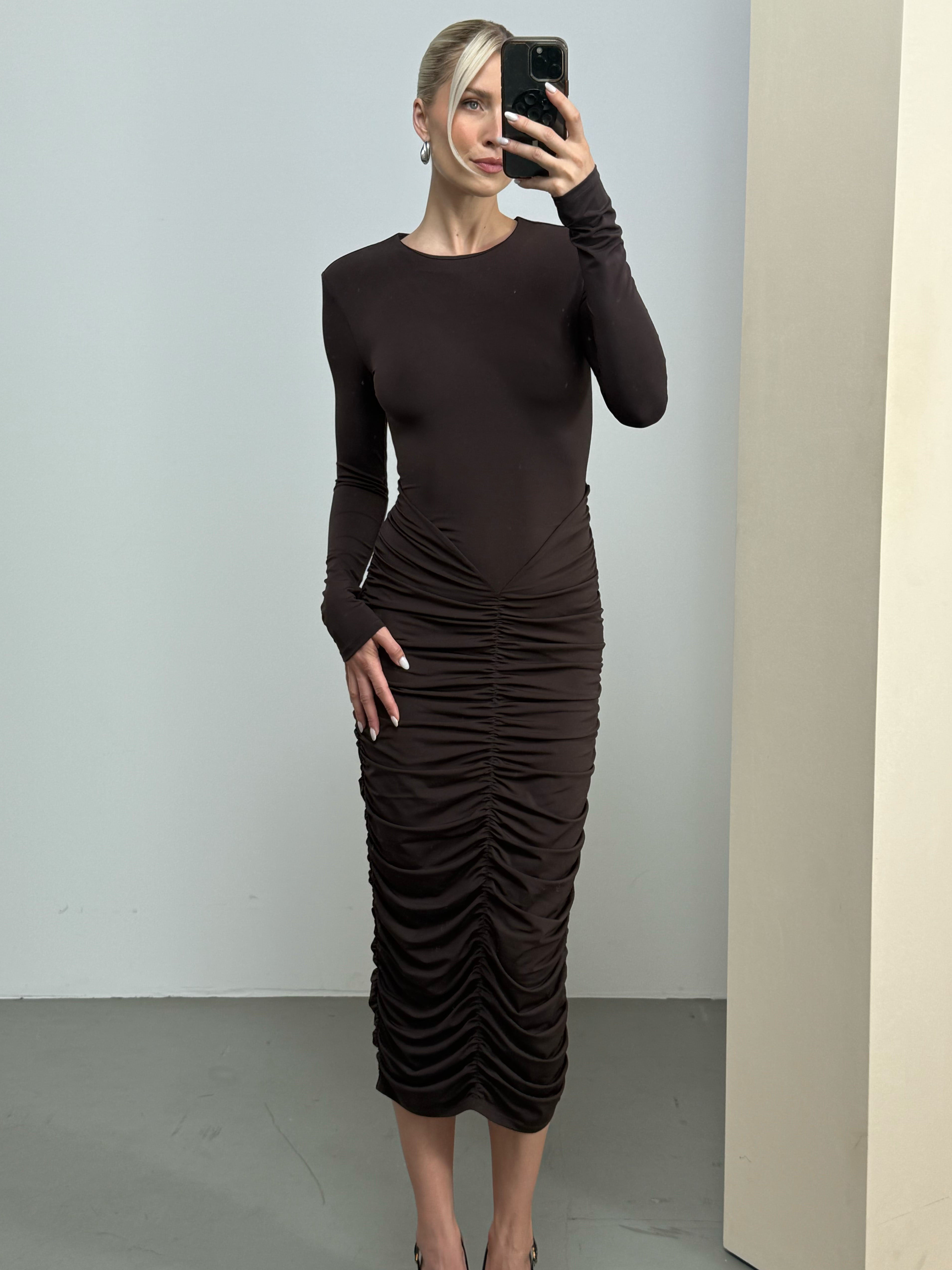 Eine Frau, die das Kleid „Johanna“ von LeGer by Lena Gercke trägt – ein figurbetontes, langärmeliges, dunkelbraunes Modell mit Rüschen –, posiert für ein Spiegel-Selfie in einem minimalistisch gestalteten Raum mit hellen Wänden und grauem Bodenbelag.