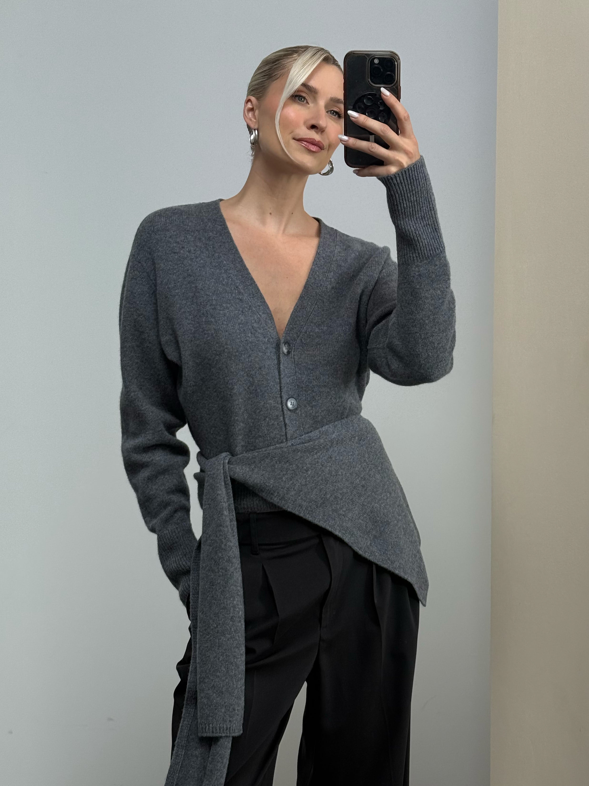 Eine Frau mit blonden Haaren, die zu einem Dutt zusammengebunden sind, macht ein Spiegel-Selfie. Sie trägt den Cardigan „Theres“ von LeGer by Lena Gercke mit stilvollem V-Ausschnitt und großen Knöpfen, dazu eine schwarze Hose mit weitem Bein. Der Hintergrund ist schlicht und minimalistisch.