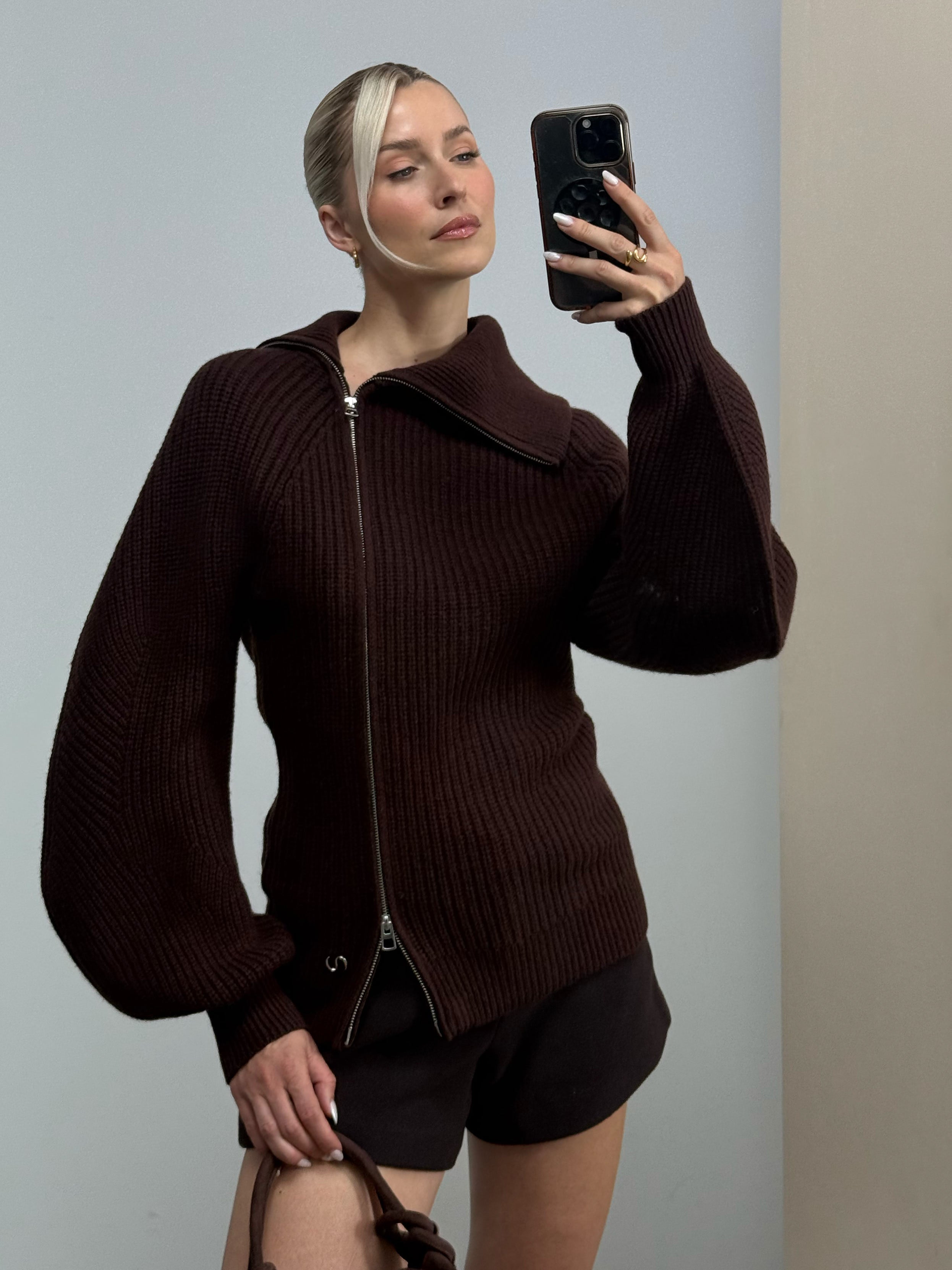 Eine Frau mit blonden Haaren, die zu einem tief sitzenden Dutt gesteckt sind, trägt den Pullover „Gioana“ von LeGer by Lena Gercke aus kuscheligem Strickmaterial, dazu Shorts, während sie ihr Handy für ein Spiegel-Selfie vor einem schlichten Hintergrund in die Hand nimmt.