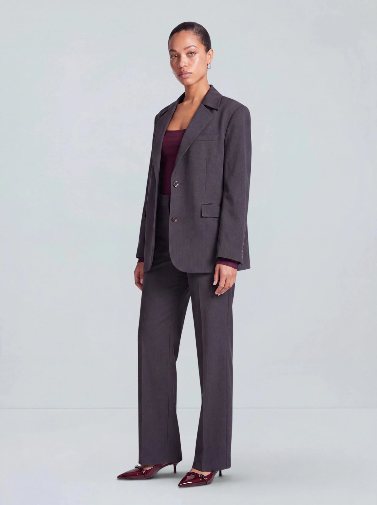 Eine Frau steht elegant mit ausgestrecktem Bein vor einem schlichten hellgrauen Hintergrund und trägt die Hose „Fergie“ von LeGer by Lena Gercke in Anthrazit, kombiniert mit einem passenden Blazer, einem weinroten Oberteil und weinroten Pumps.