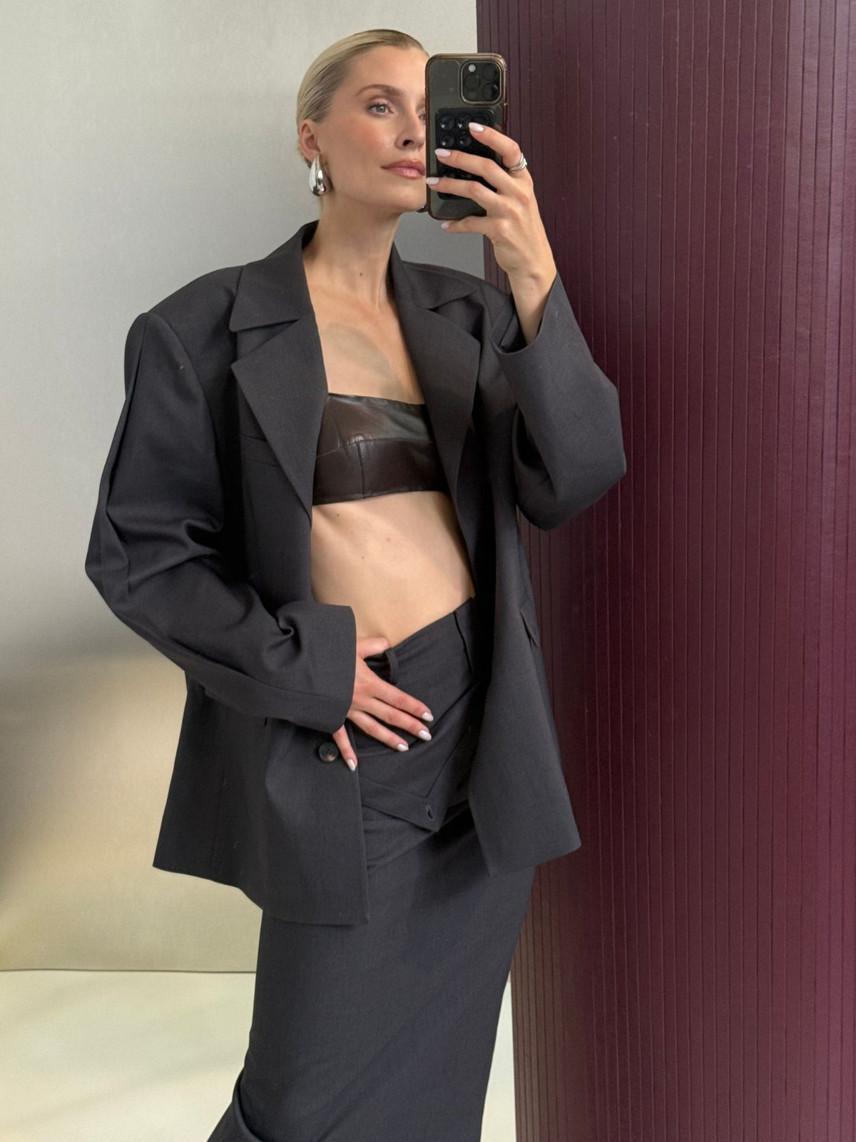 Eine Person macht ein Spiegel-Selfie und trägt dabei den Blazer „Allie“ von LeGer by Lena Gercke in Anthrazit, der in einer Oversize-Passform über einem schwarzen Bandeau-Top und einem dazu passenden dunklen Rock kombiniert ist. Die Haare sind nach hinten gesteckt, während die Person vor einer strukturierten, burgunderroten Wand steht.