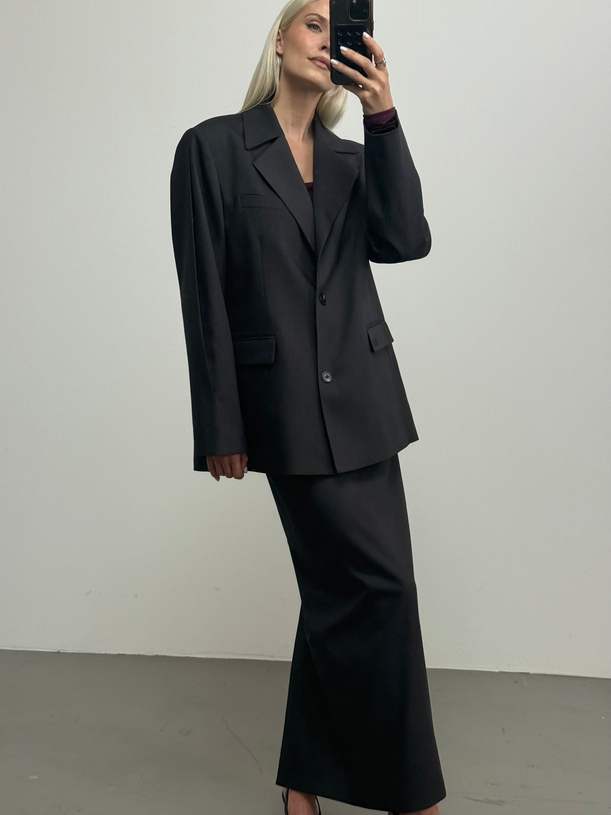 Eine Person mit langen blonden Haaren trägt das „Allie“-Set von LeGer by Lena Gercke – einen Oversize-Blazer und einen dazu passenden Bleistiftrock in Anthrazit –, steht in einem Raum und macht vor einer schlichten weißen Wand ein Spiegel-Selfie.