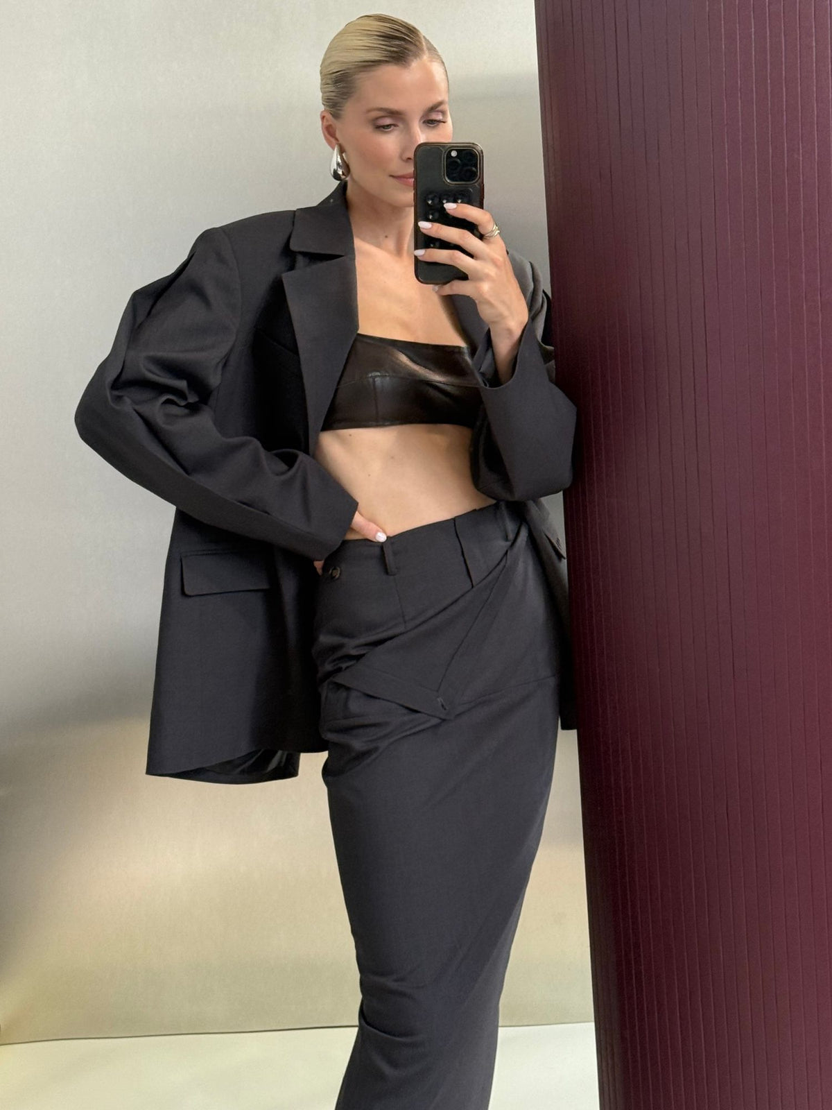 Eine Person in einem schwarzen Oversize-Blazer, einem schwarzen Leder-Bandeau-Top und dem Rock „Allie“ von LeGer by Lena Gercke macht ein Spiegel-Selfie, während sie vor einer burgunderroten Wand steht.