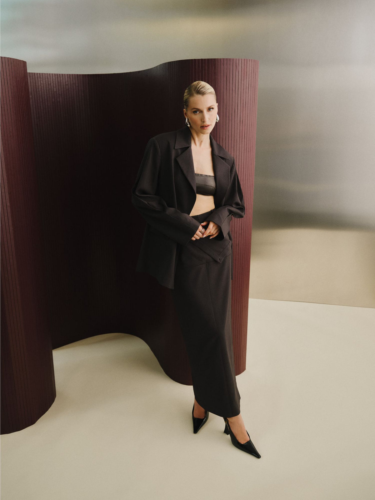 Eine Frau, die den Rock „Allie“ von LeGer by Lena Gercke trägt – einen hoch taillierten, knöchellangen Bleistiftrock – kombiniert diesen mit einem dunklen Oversize-Blazer und einem Bandeau-Top, während sie vor einer geschwungenen, burgunderroten Trennwand auf einem cremefarbenen Boden steht. Abgerundet wird der Look durch spitze schwarze Pumps und Creolen.