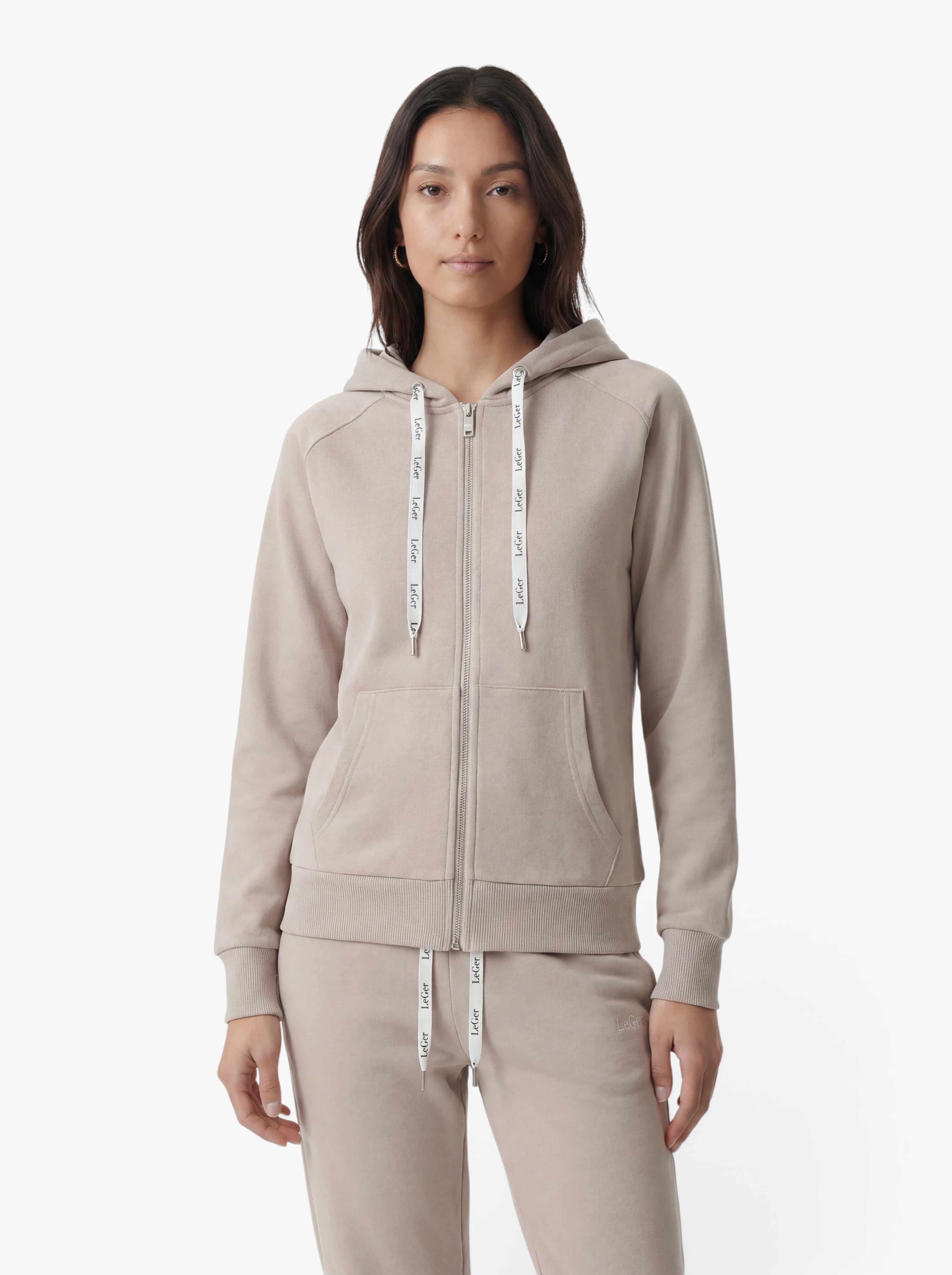 Eine Person trägt den Hoodie „Phoenix“ aus der LeGer by Lena Gercke-Kollektion in Hellbeige, dazu eine passende Jogginghose, und steht vor einem schlichten weißen Hintergrund. Der Hoodie wurde für optimalen Komfort bei sportlichen Aktivitäten entworfen und verfügt über markante weiße Kordeln mit Textdruck.