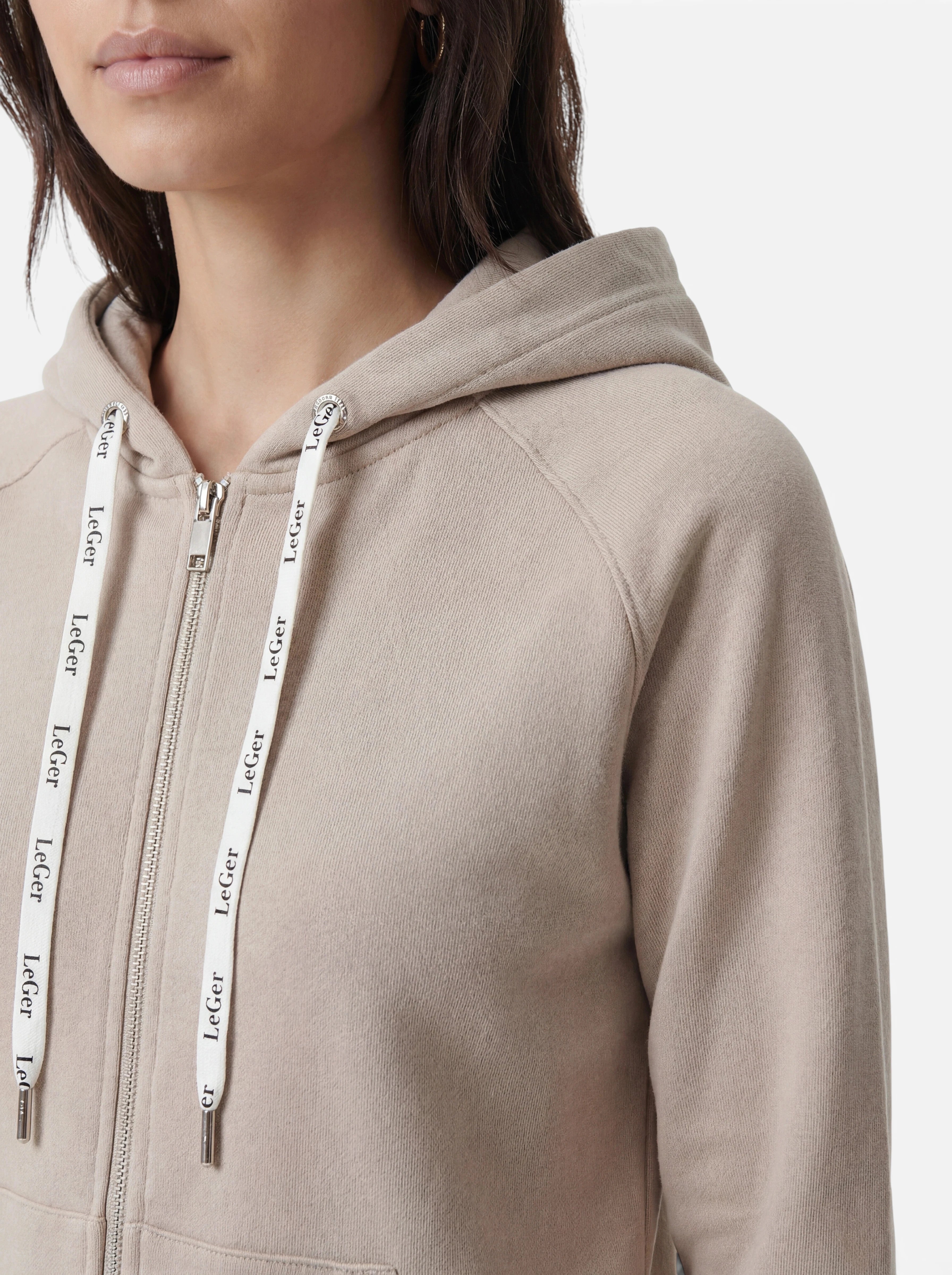 Eine Person, die den „Phoenix“-Hoodie von LeGer by Lena Gercke trägt – einen beigen Hoodie mit Reißverschluss und weißen Kordeln, auf denen „LeGer“ aufgedruckt ist –, ist vor weißem Hintergrund von den Schultern bis zur Körpermitte zu sehen. Dieser Hoodie bietet die perfekte Mischung aus Komfort und Stil für sportliche Aktivitäten.