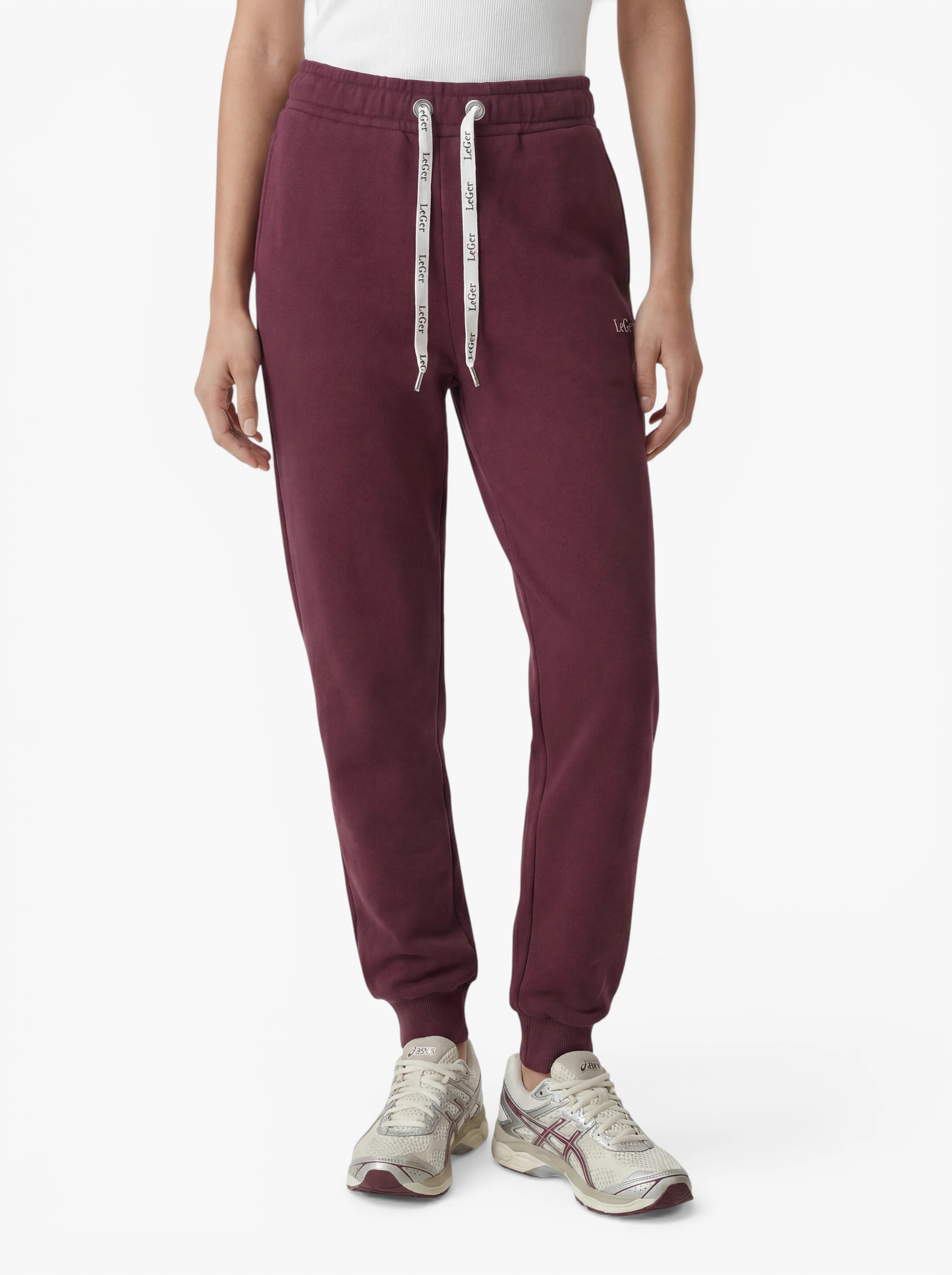 Eine Person trägt die burgunderrote „Nela“-Hose von LeGer by Lena Gercke – eine Jogginghose, die vom eleganten Hosenstil inspiriert ist und über weiße Kordelzüge verfügt. Kombiniert mit einem weißen Oberteil und beigen Sneakern ist das Bild so beschnitten, dass nur der Körper von den Schultern abwärts vor einem schlichten weißen Hintergrund zu sehen ist, wodurch die schmeichelhafte Silhouette perfekt zur Geltung kommt.