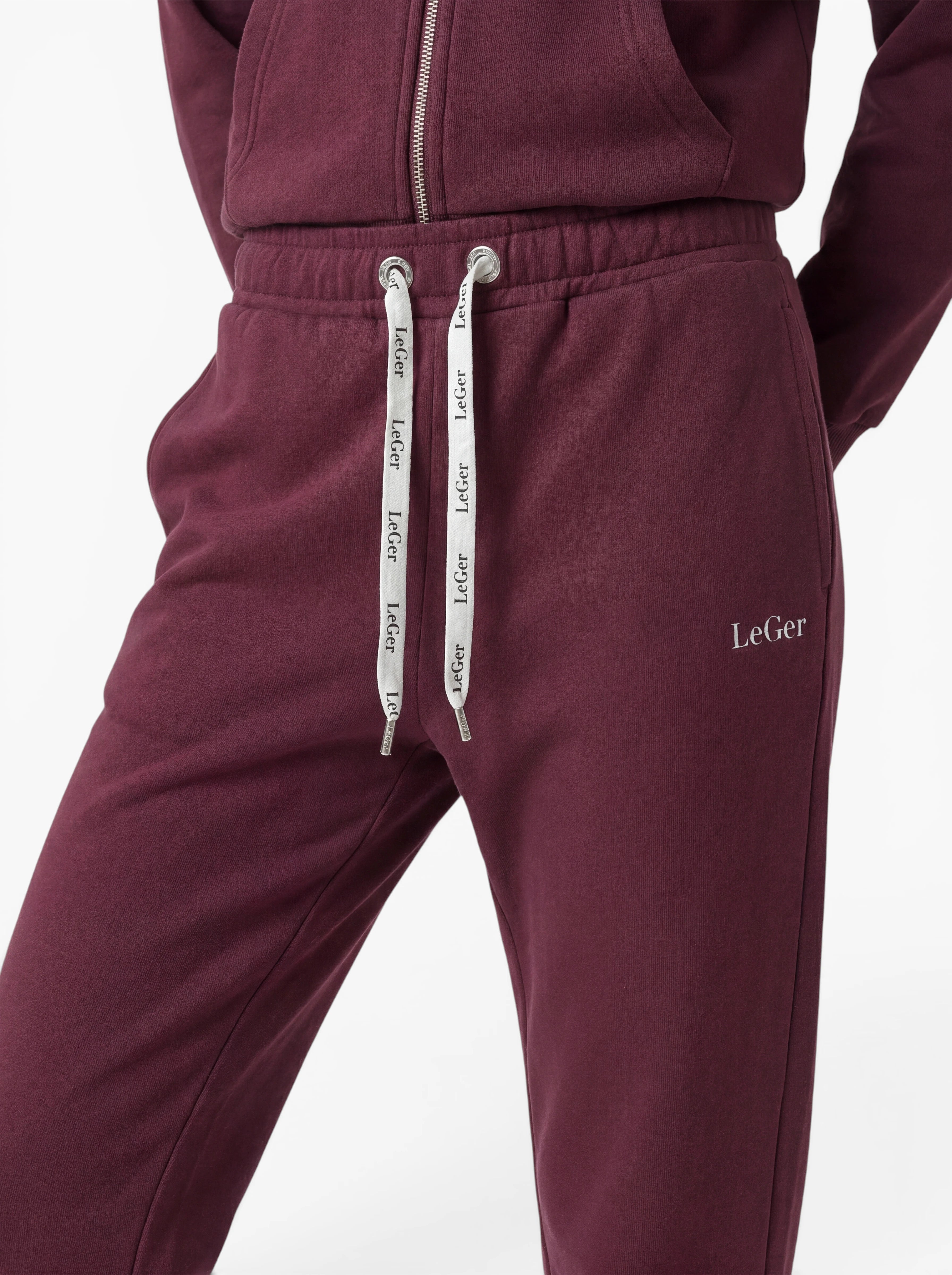 Eine Person trägt die Hose „Nela“ von LeGer by Lena Gercke – eine burgunderrote Jogginghose mit weißen Kordeln, die eine schmeichelhafte Silhouette zaubert. Der dazu passende Kapuzenpulli mit Reißverschluss ist teilweise zu sehen, und die Hände ruhen auf den Hüften.